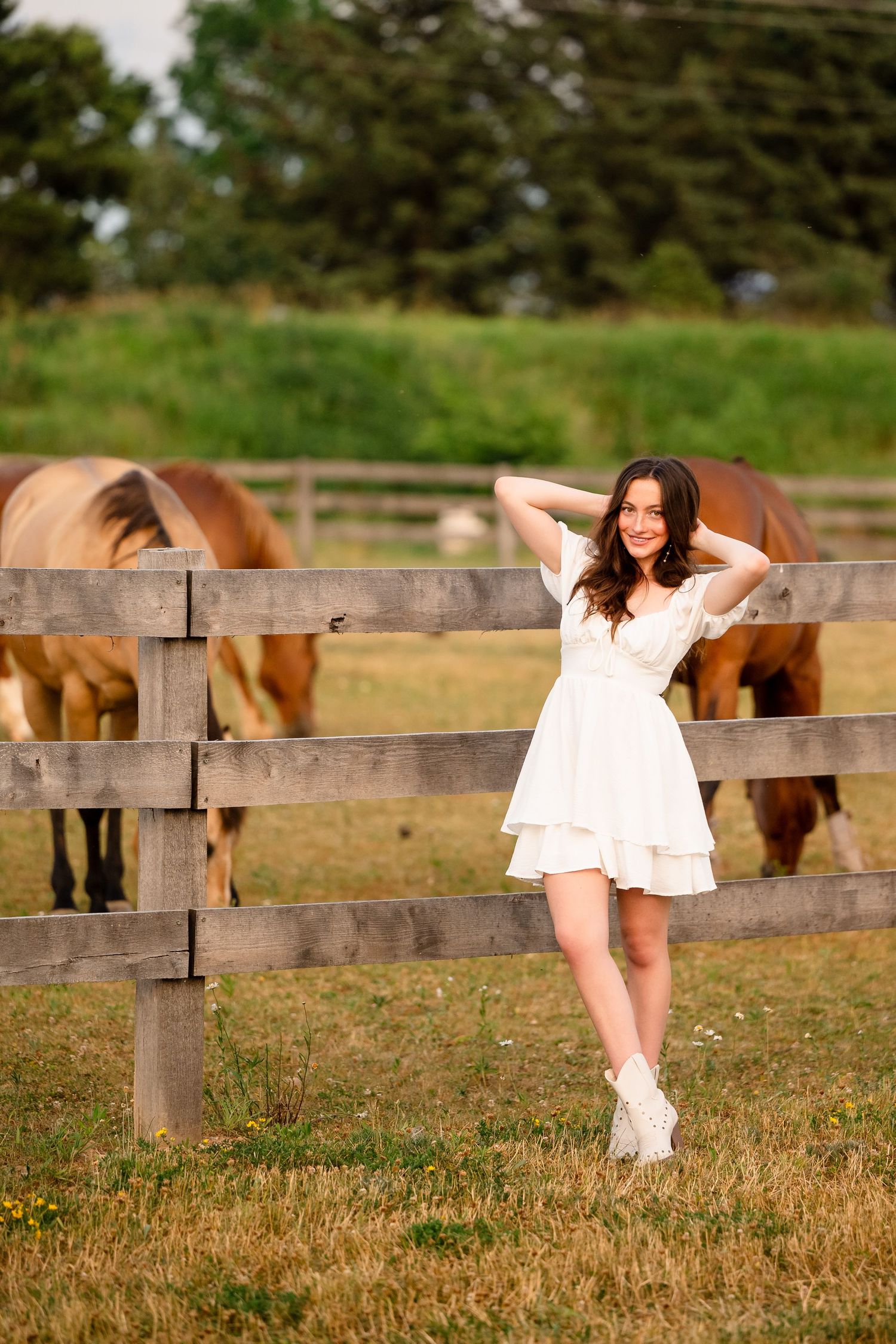 A woman in a white dress poses playfully near a wooden fence with horses grazing in the background.