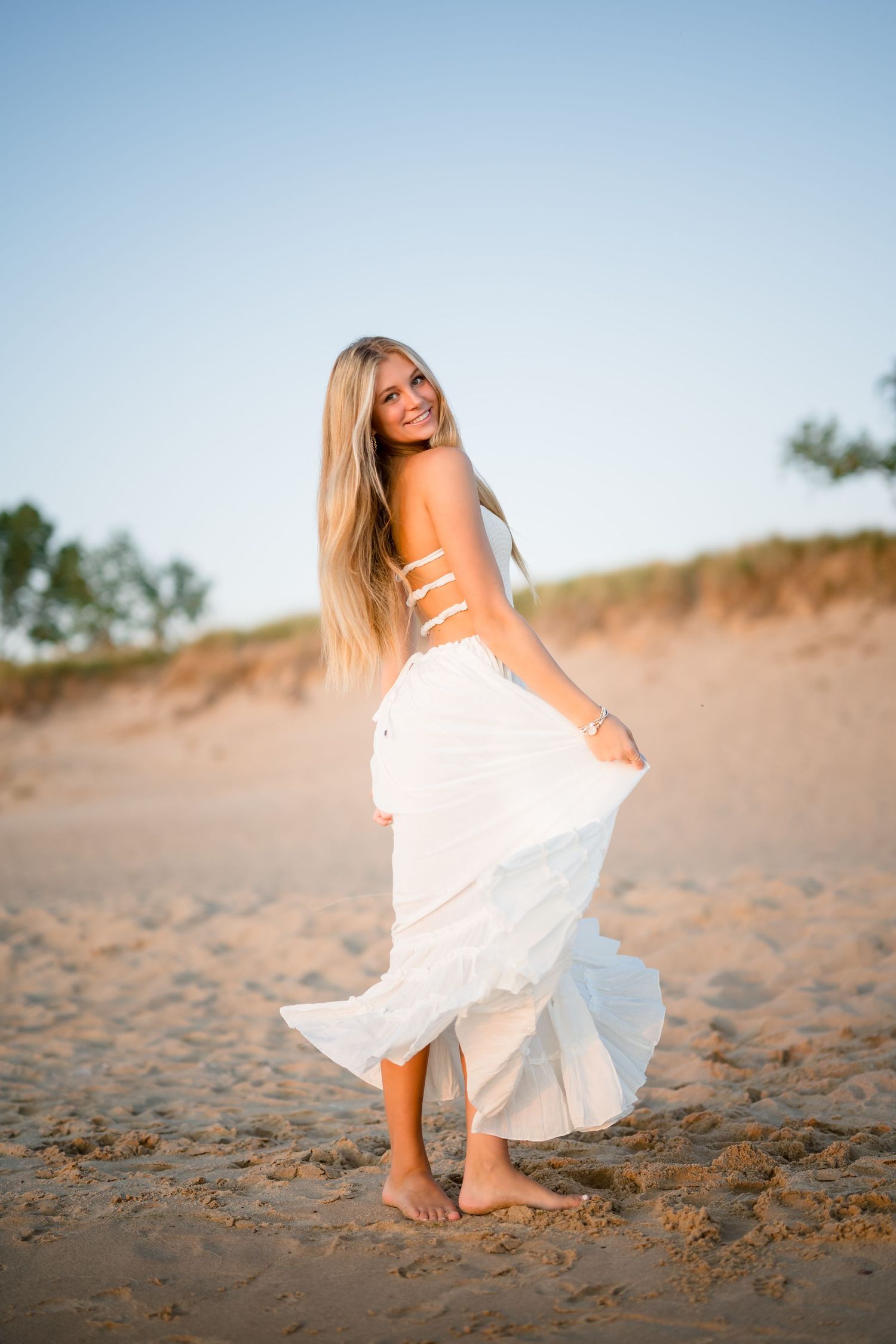 Girl in white dress poses playfully on sandy beach at sunset during a senior photo session at Lake Michigan.