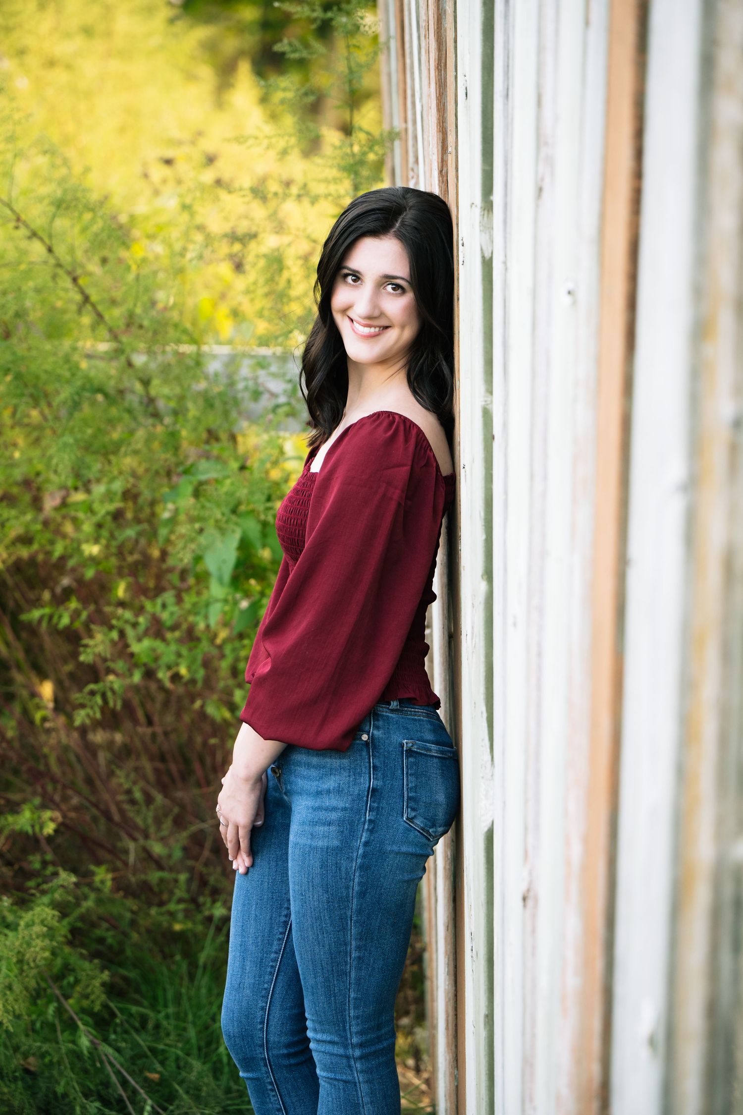 Casual outdoor portrait against a natural yellow-green background wearing a burgundy sweater and blue jeans.