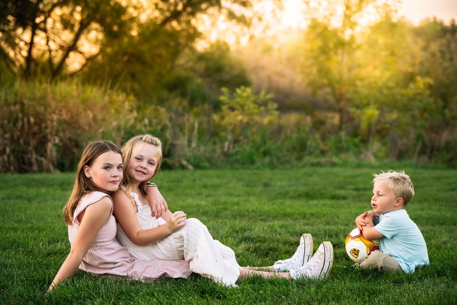 Three children sitting on green grass at sunset, two girls in pink and white dresses and a boy holding a teddy bear.