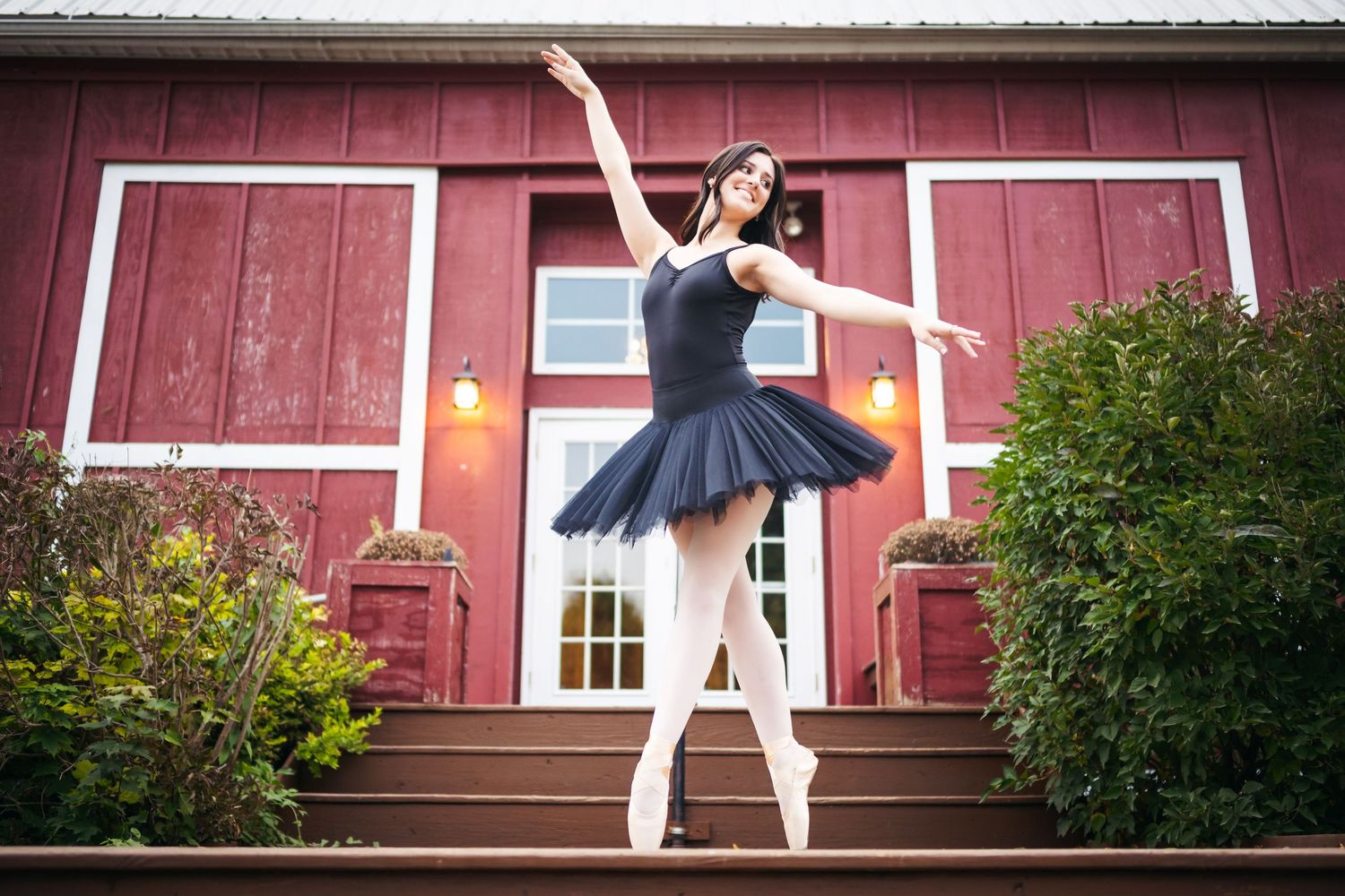 Ballet dancer in a navy tutu performs graceful poses against a rustic red barn backdrop.