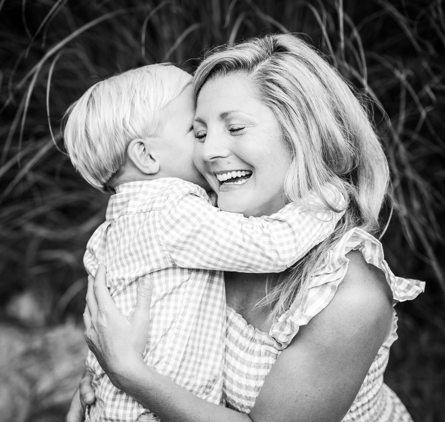 Black and white photo of a laughing woman hugging a young blonde boy who kisses her cheek outdoors.