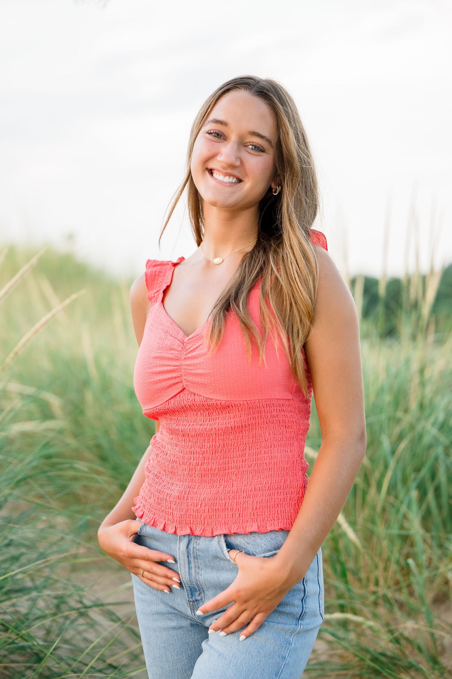 A person in a coral tank top and jeans poses outdoors in tall grass during a summer photoshoot.