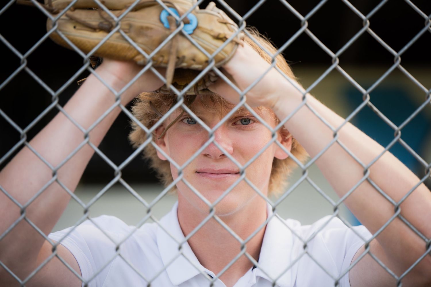 A senior guy leans against a chain-link fence while wearing a white shirt and looking contemplatively into the distance.