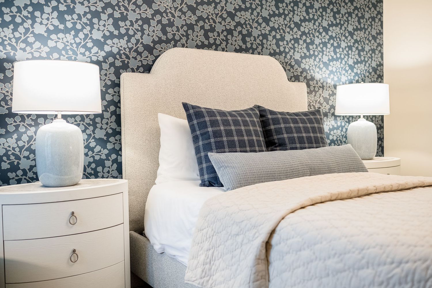 Modern bedroom interior with gray wallpaper, white bedding, and plaid accent pillows on a comfortable bed.