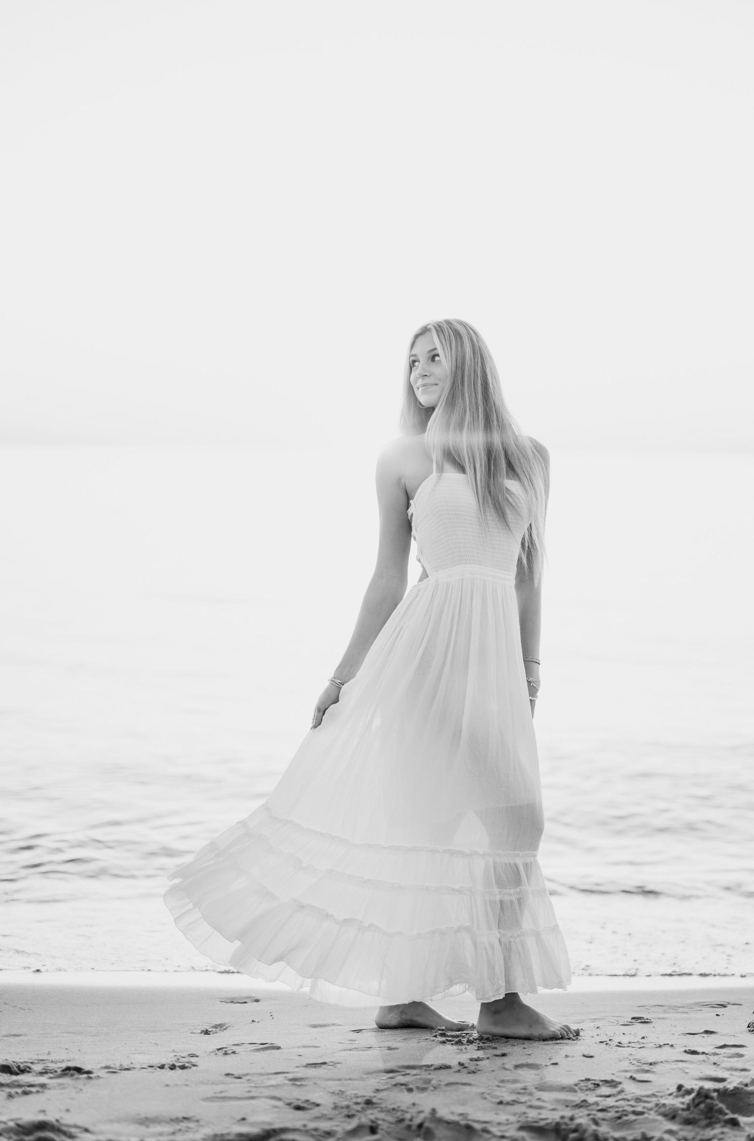Black and white photo of a young woman in a flowing white maxi dress standing barefoot on a sandy beach.
