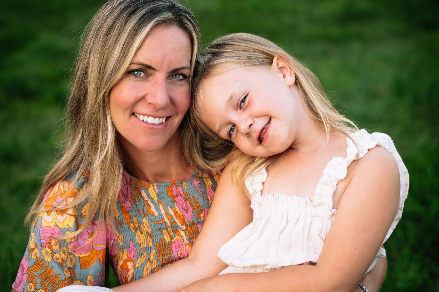 Mom and daughter share a warm moment outdoors against a green background, wearing colorful and white clothing during family portrait session