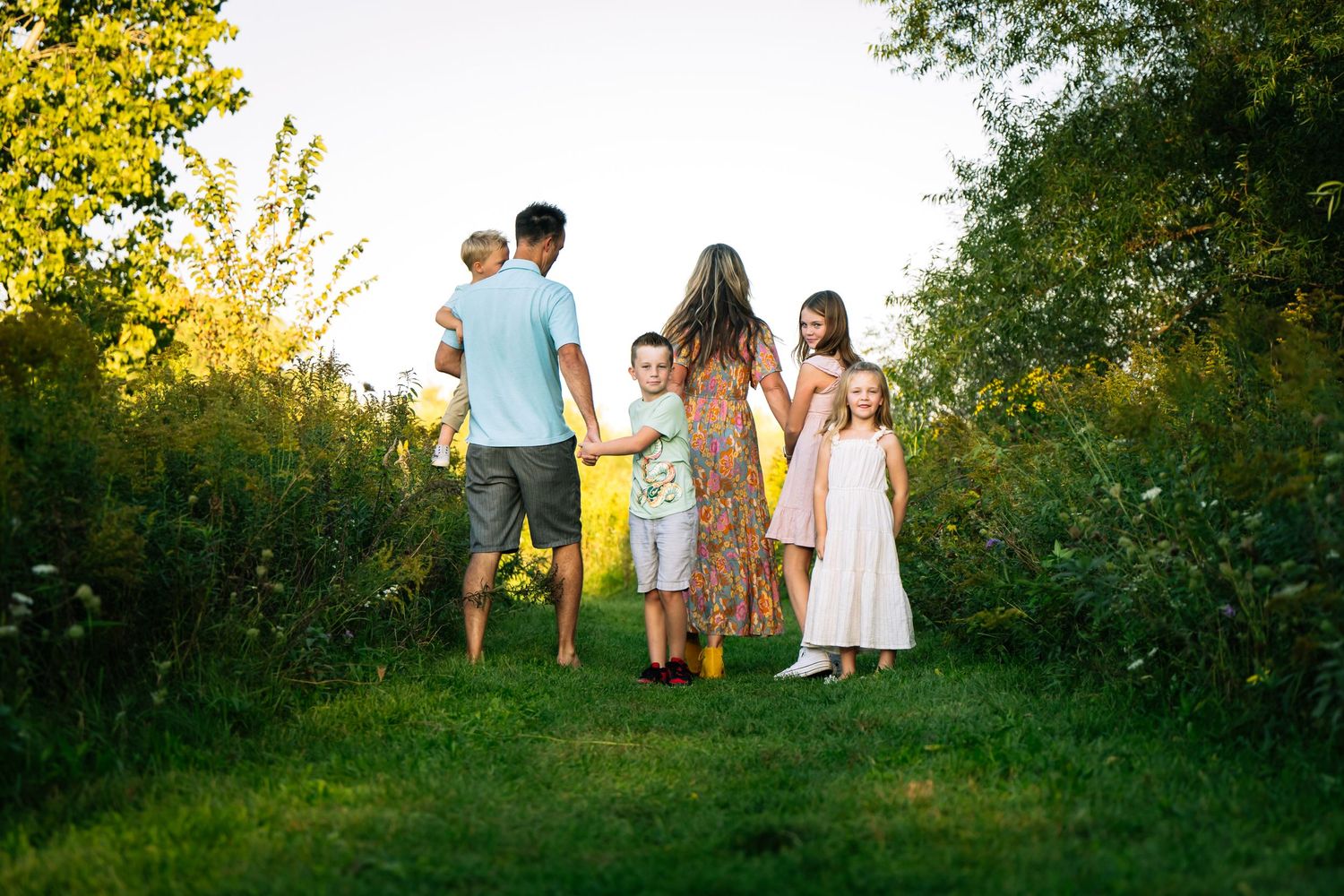 A family walks together along a sunlit nature path surrounded by lush greenery on a summer evening.