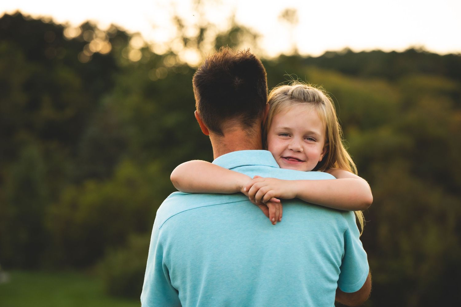 A child in a light blue shirt rests their head on someone's shoulder during golden hour outdoors.