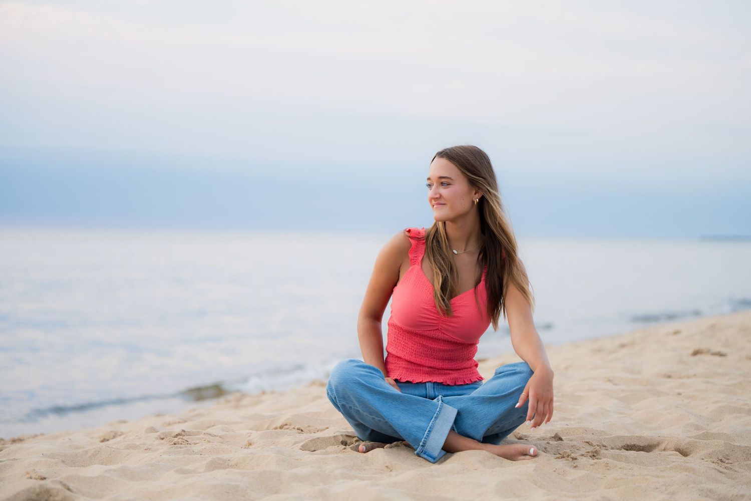 A senior girl  in a coral top and jeans sits casually on a sandy beach with a bright sky in the background.