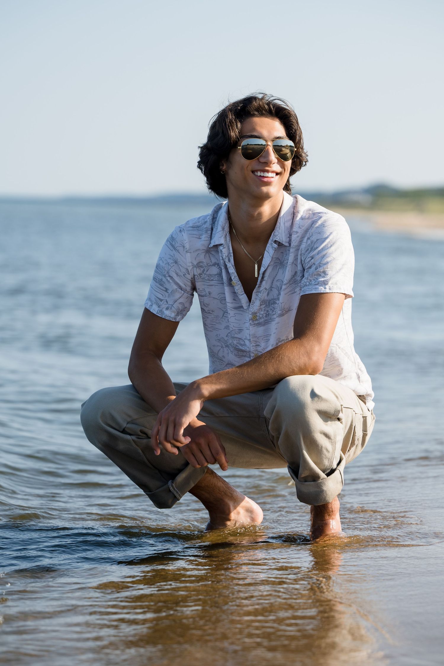 Person in white shirt and khaki pants crouches at the edge of the ocean water on a sunny beach day.