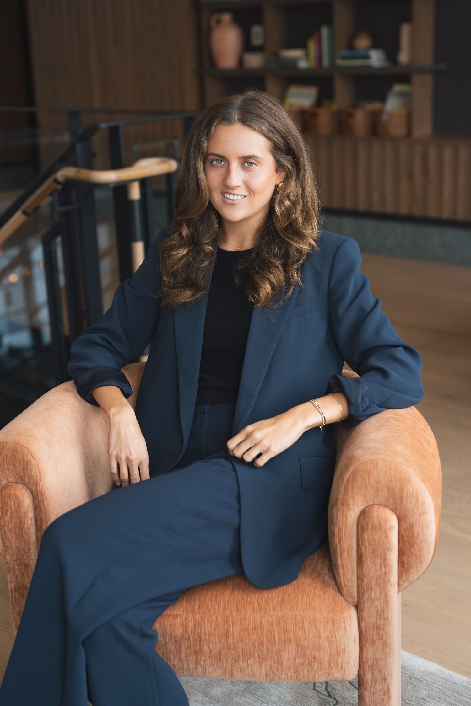 Professional wearing a navy blue business suit sits relaxed in a tan armchair in a modern office setting.
