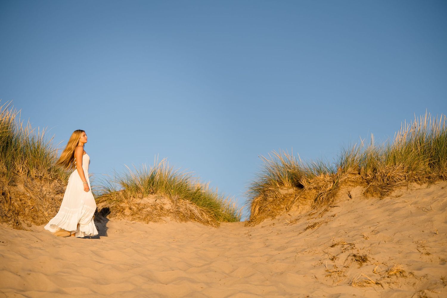 A white wedding dress flows elegantly against sandy dunes at golden hour on a beach.