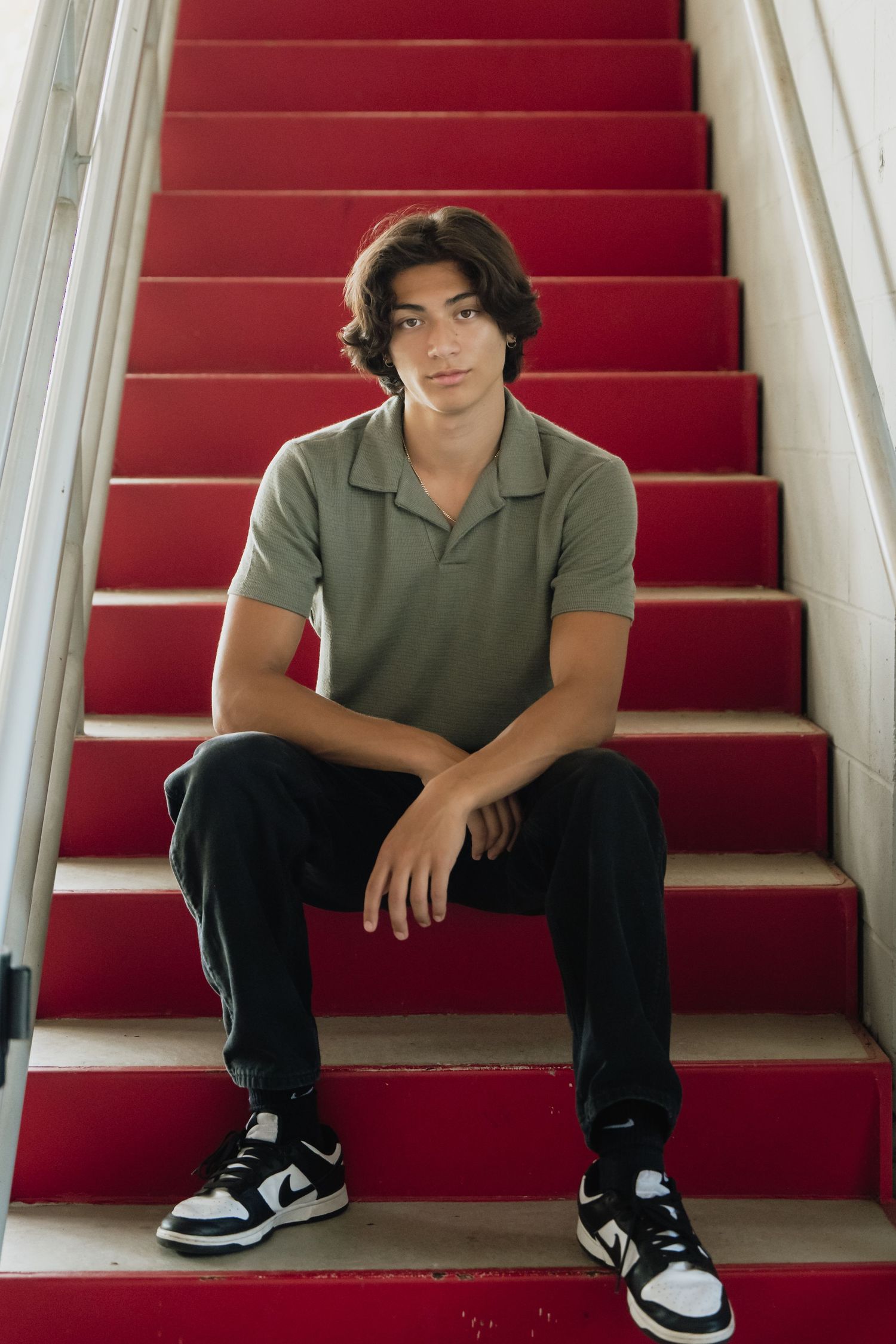 A person in a green polo shirt and black pants sits on red carpeted stairs wearing black and white sneakers.