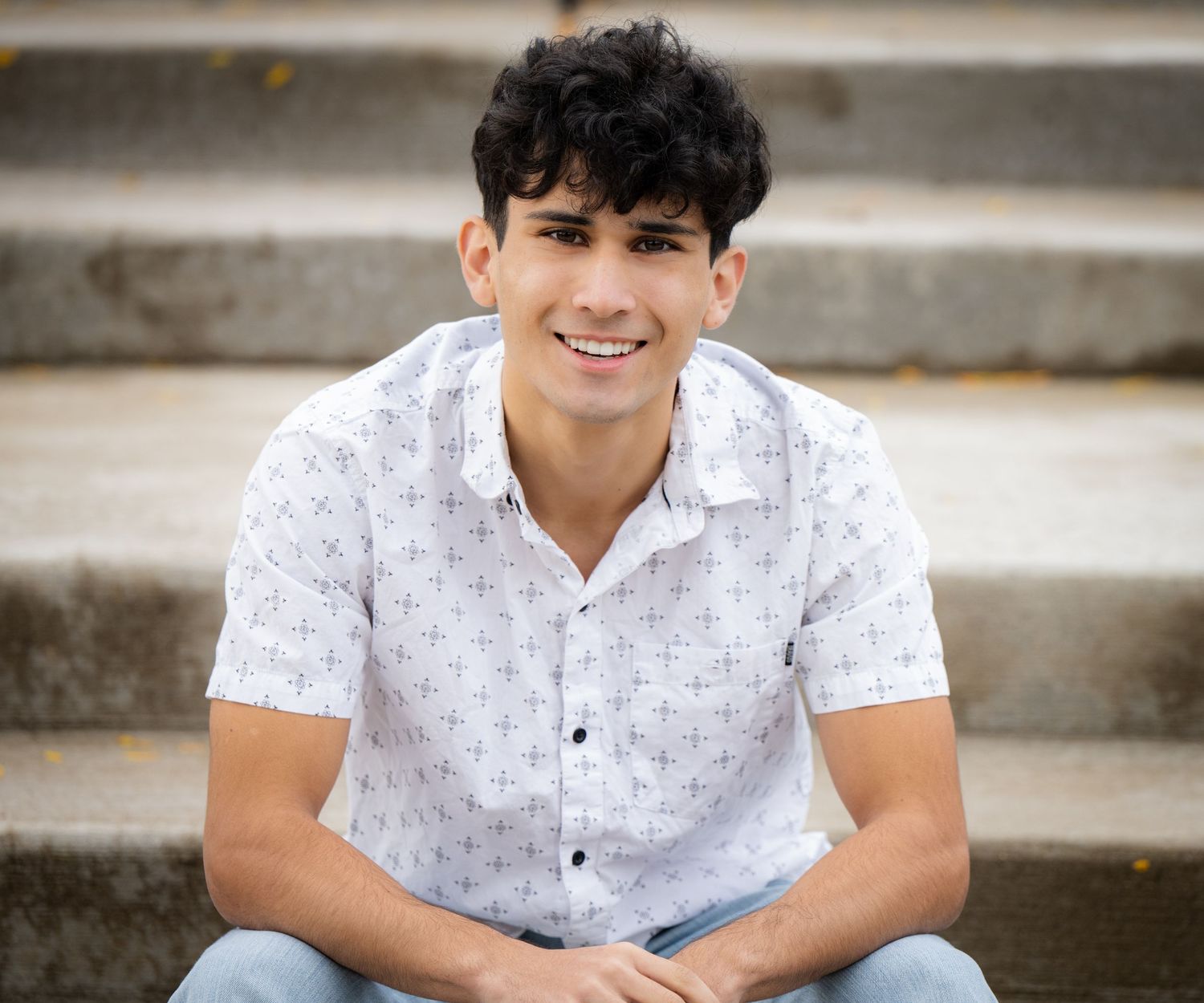 A smiling young man in a white patterned button-up shirt sits on stone steps outdoors.