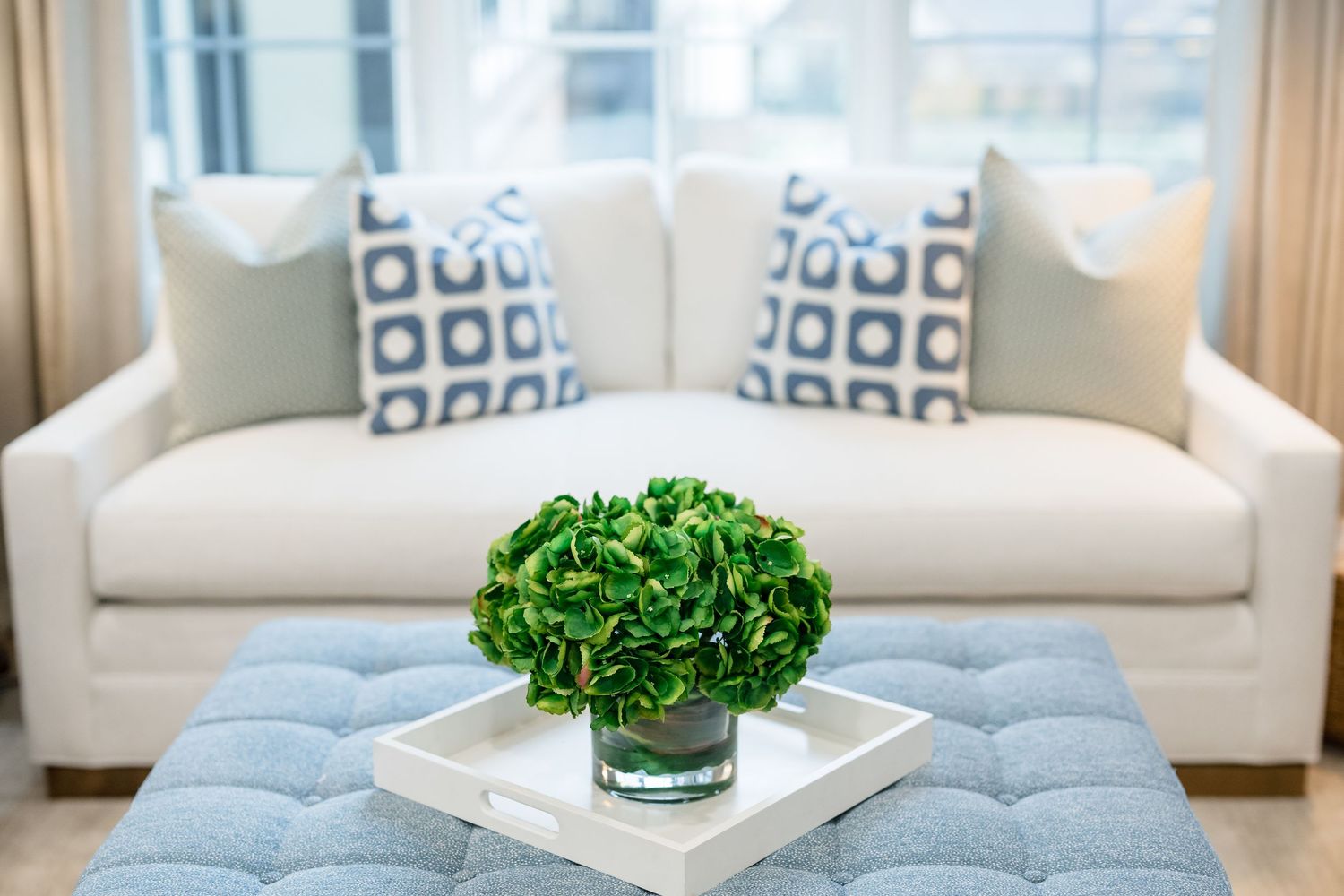 Modern coastal living room features white sofas, blue geometric pillows, and natural greenery accents.