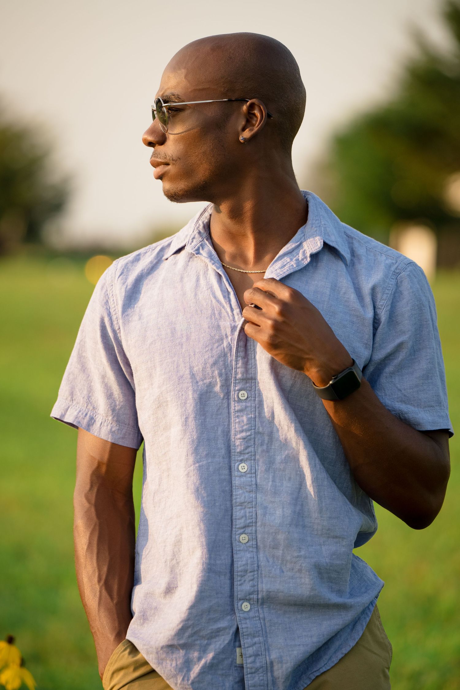 Senior guy in a light blue button-down shirt poses thoughtfully against a blurred natural outdoor background.