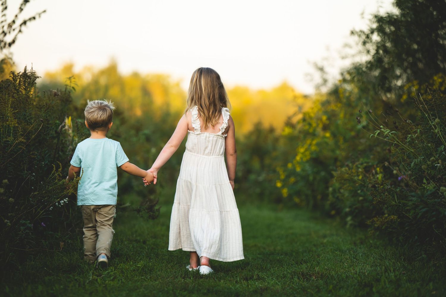 Two children holding hands while walking on a grassy path at sunset with golden light filtering through trees.