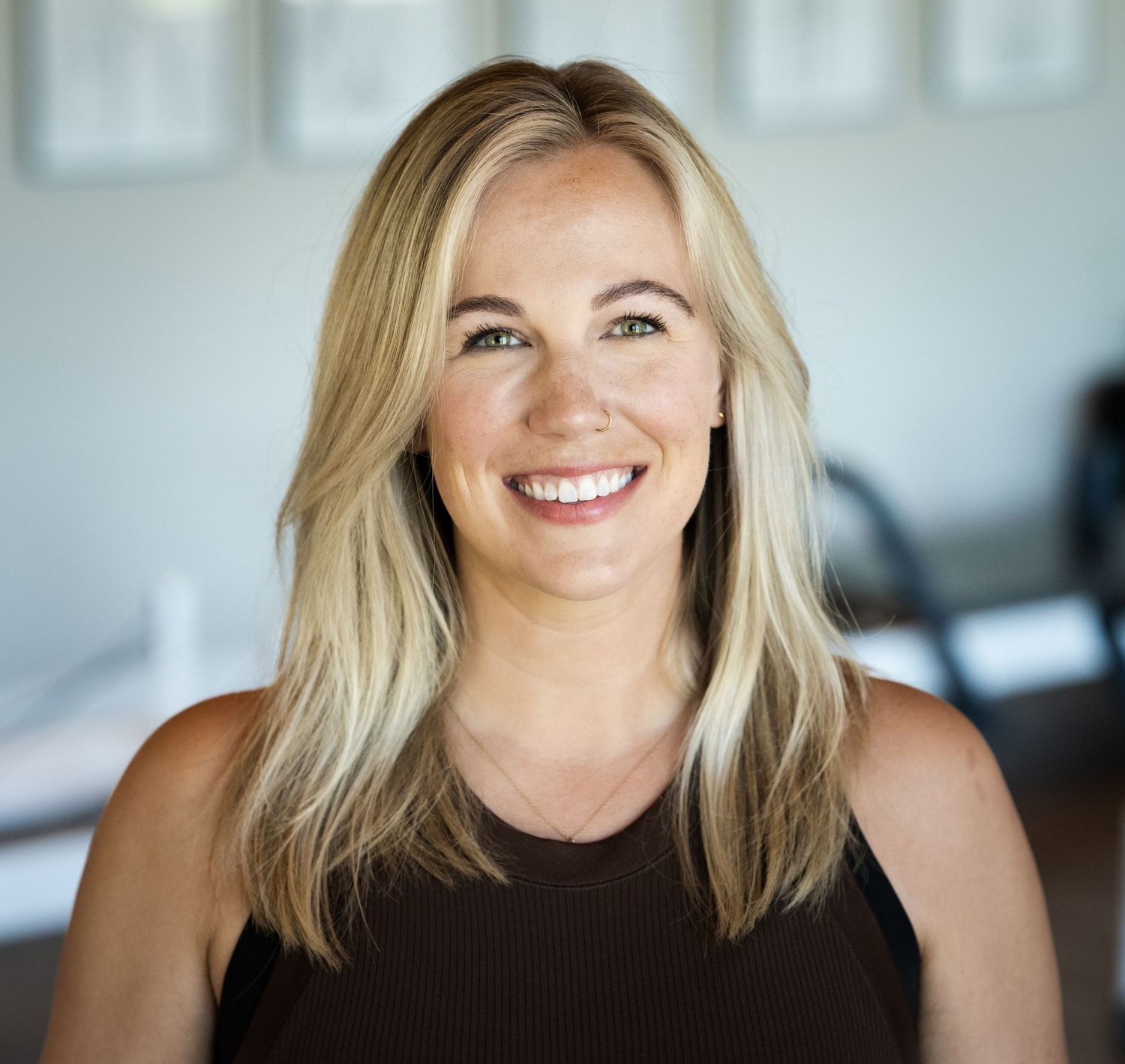 Professional headshot of a blonde woman in a black sleeveless top smiling warmly against a bright interior background.