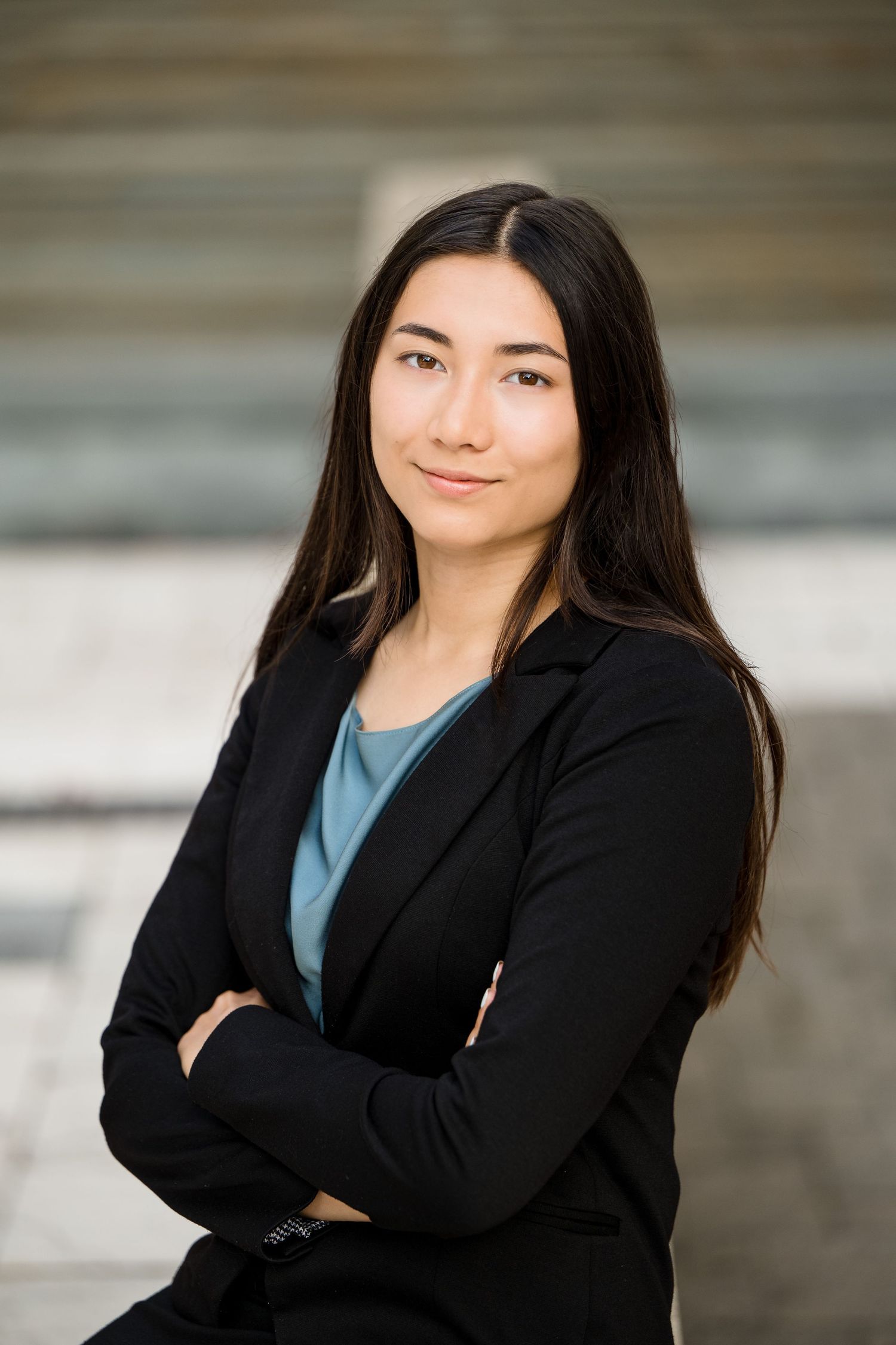 Professional headshot of a person in a black blazer and blue top with long dark hair against a blurred background.