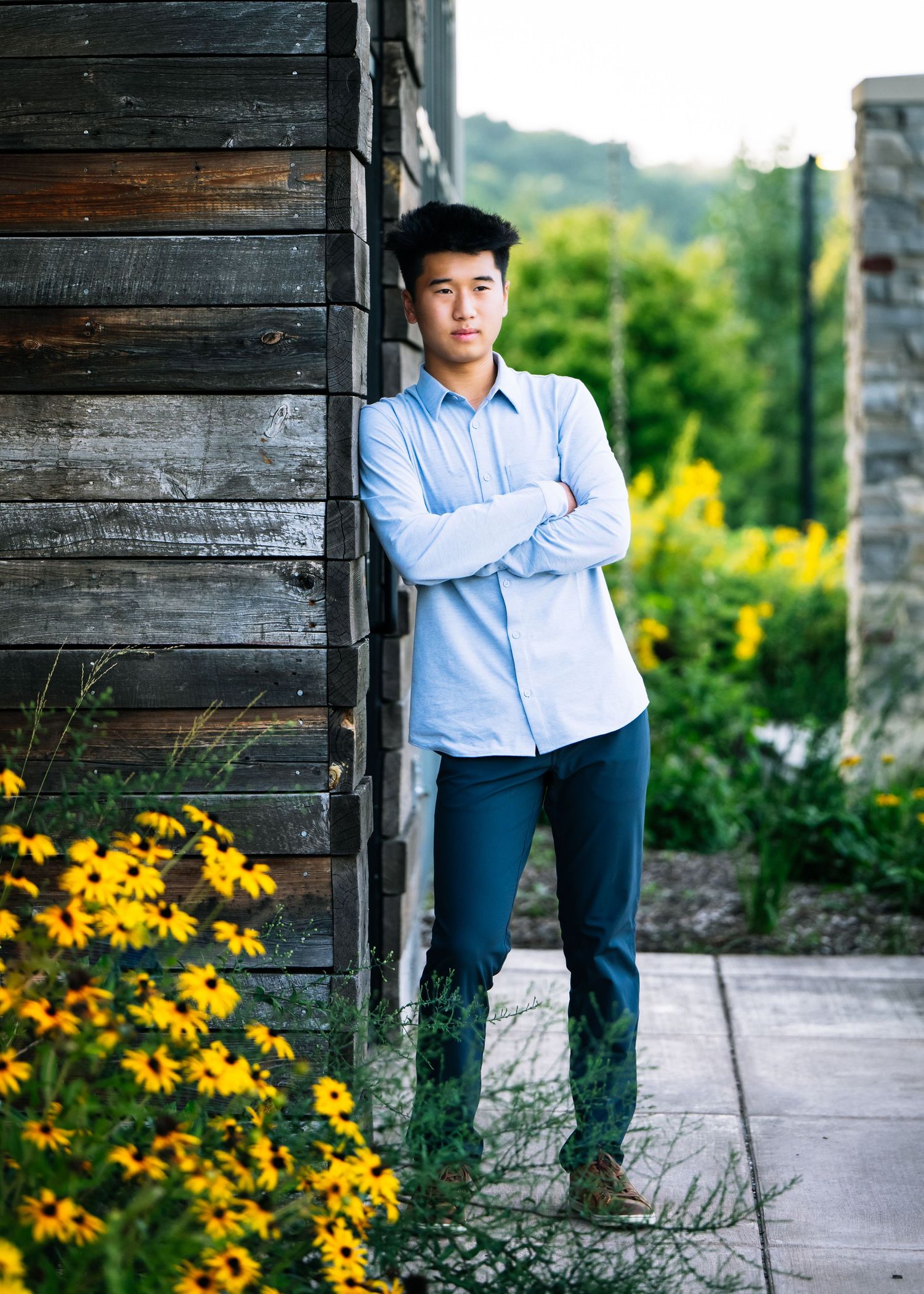 Person in blue shirt and dark pants poses against rustic wooden wall with vibrant yellow flowers in garden setting.