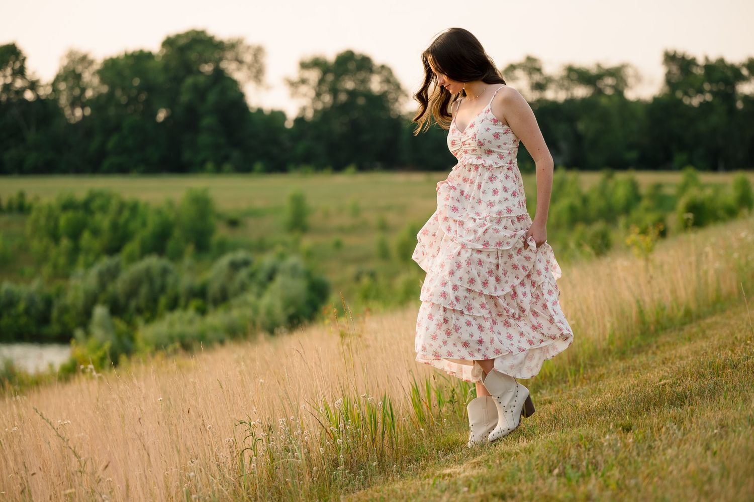 A young woman in a floral sundress and white boots walks through a lush green meadow at golden hour.
