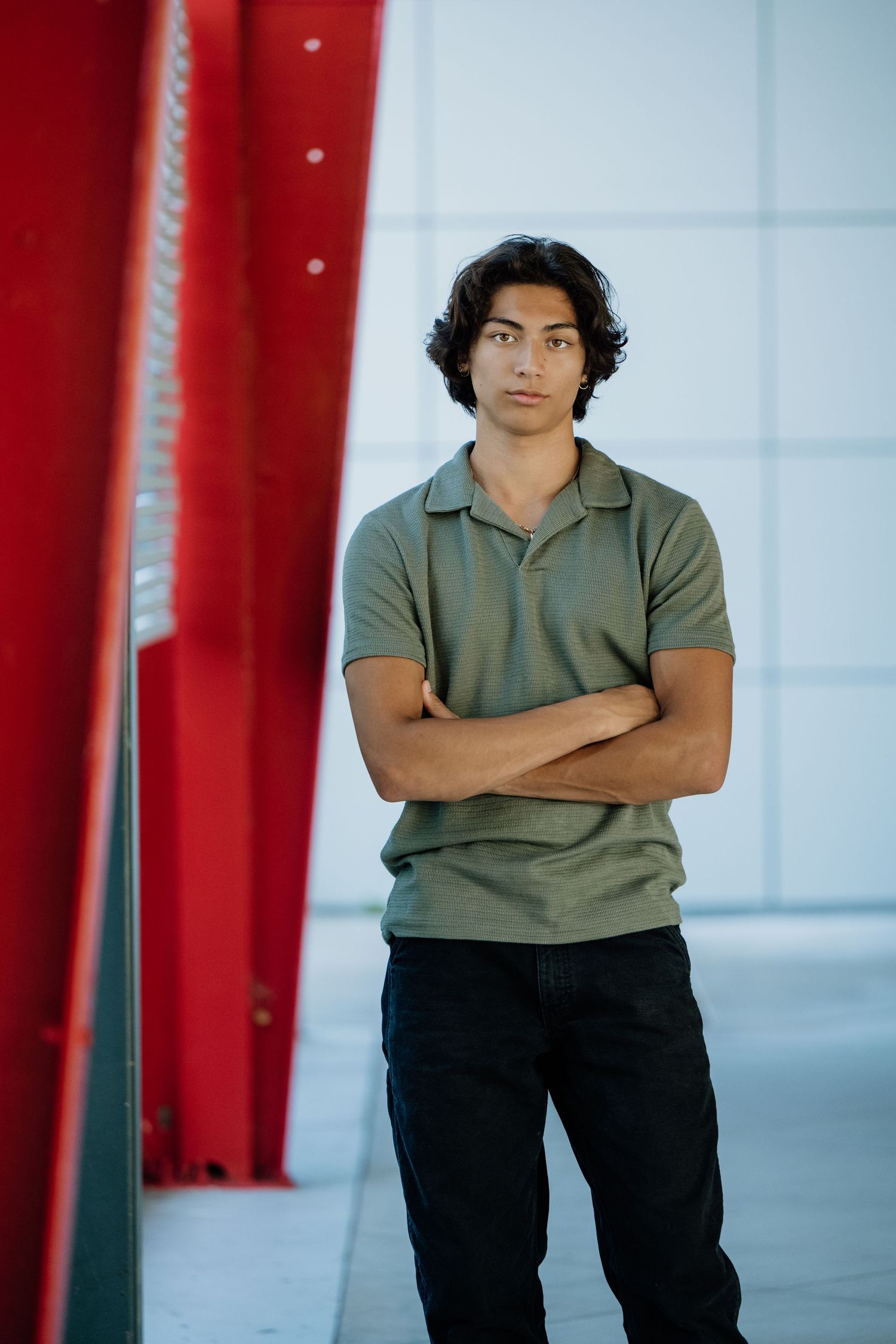 Young man with crossed arms in olive polo shirt stands confidently beside a red structure in a modern setting.