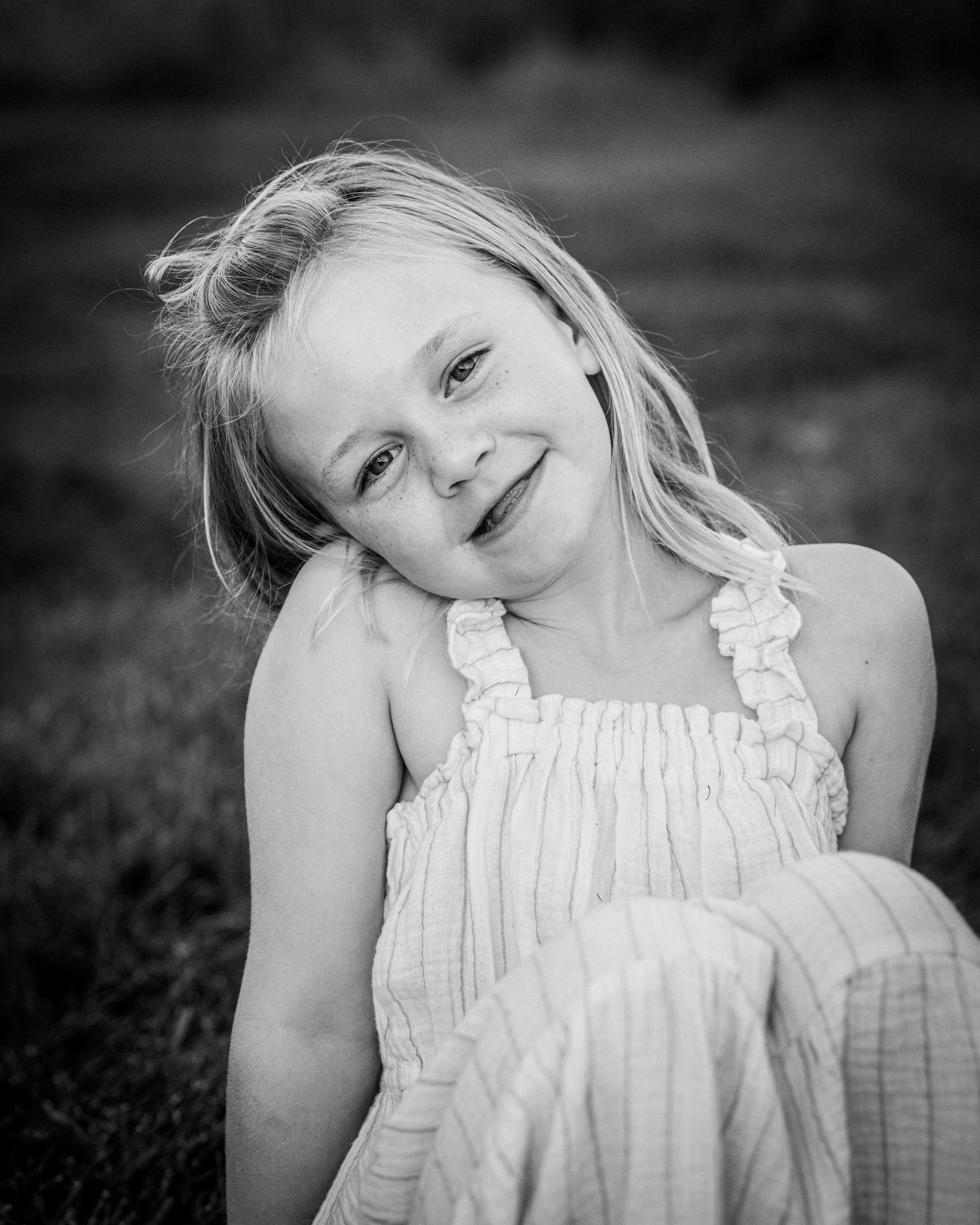 Black and white portrait of a child in a white summer dress tilting their head and smiling at the camera.