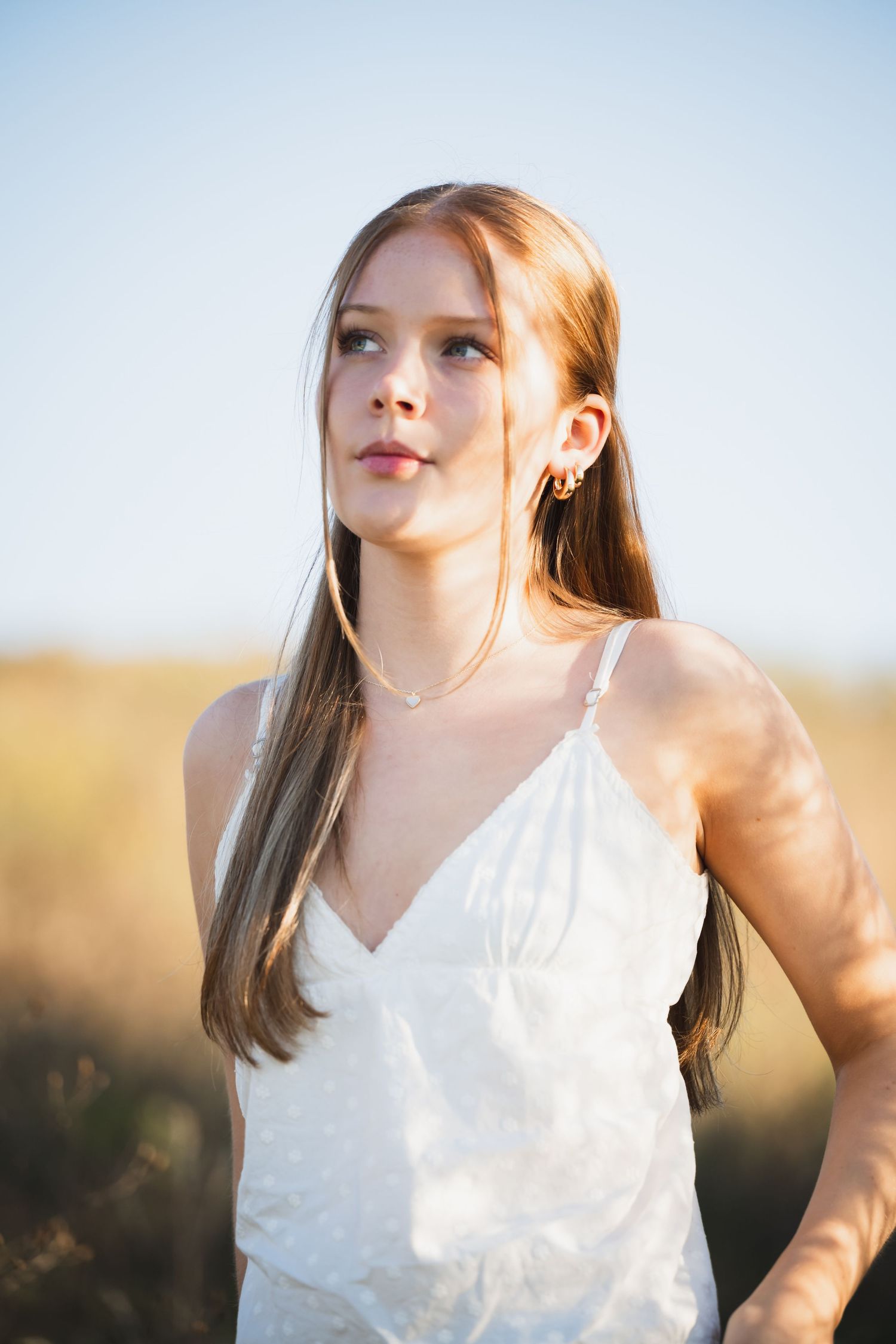 A senior girl in a white top poses outdoors during golden hour with soft lighting and natural scenery in the background.