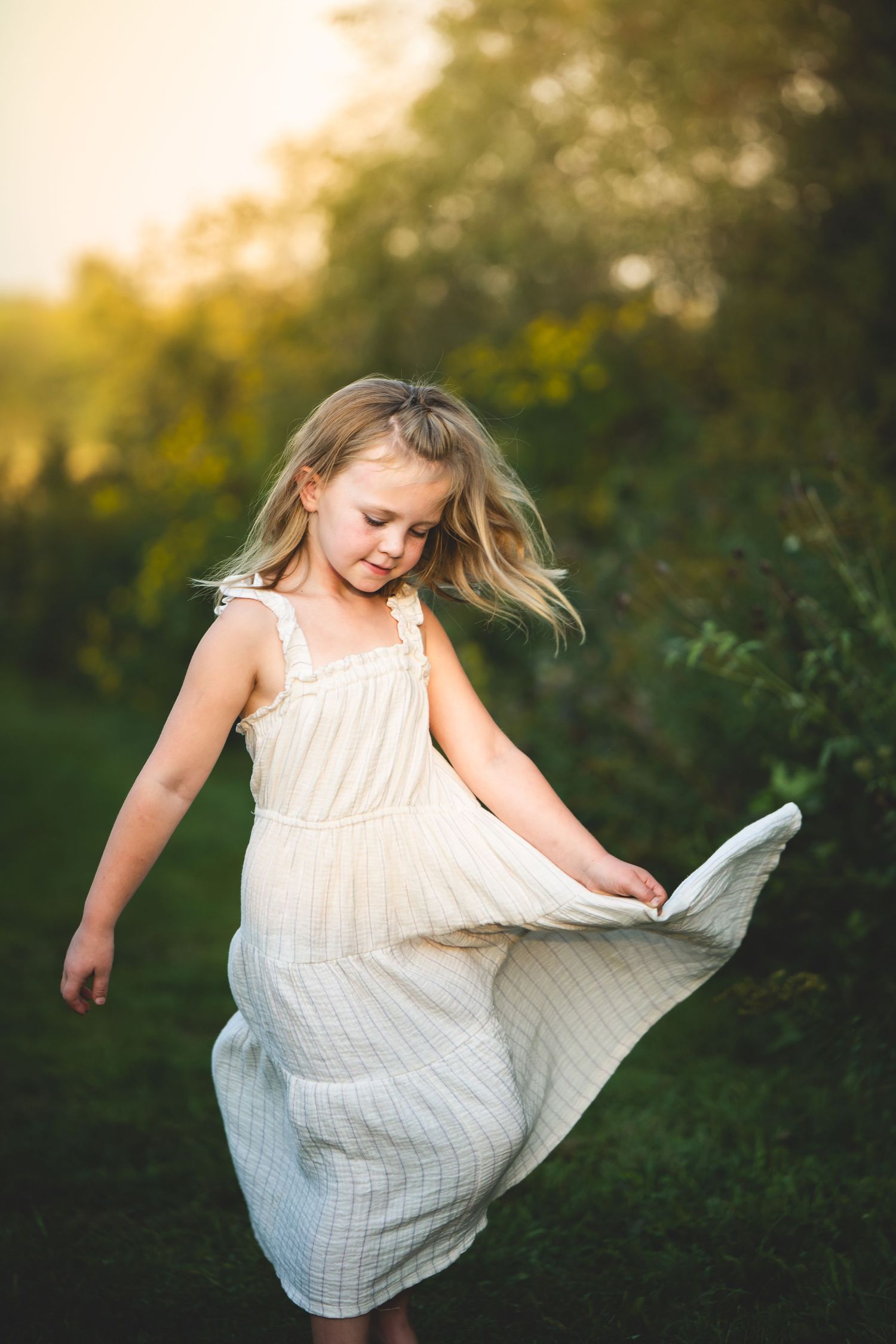 A young girl in a white summer dress twirls and dances in a sunlit garden during golden hour family photo session.