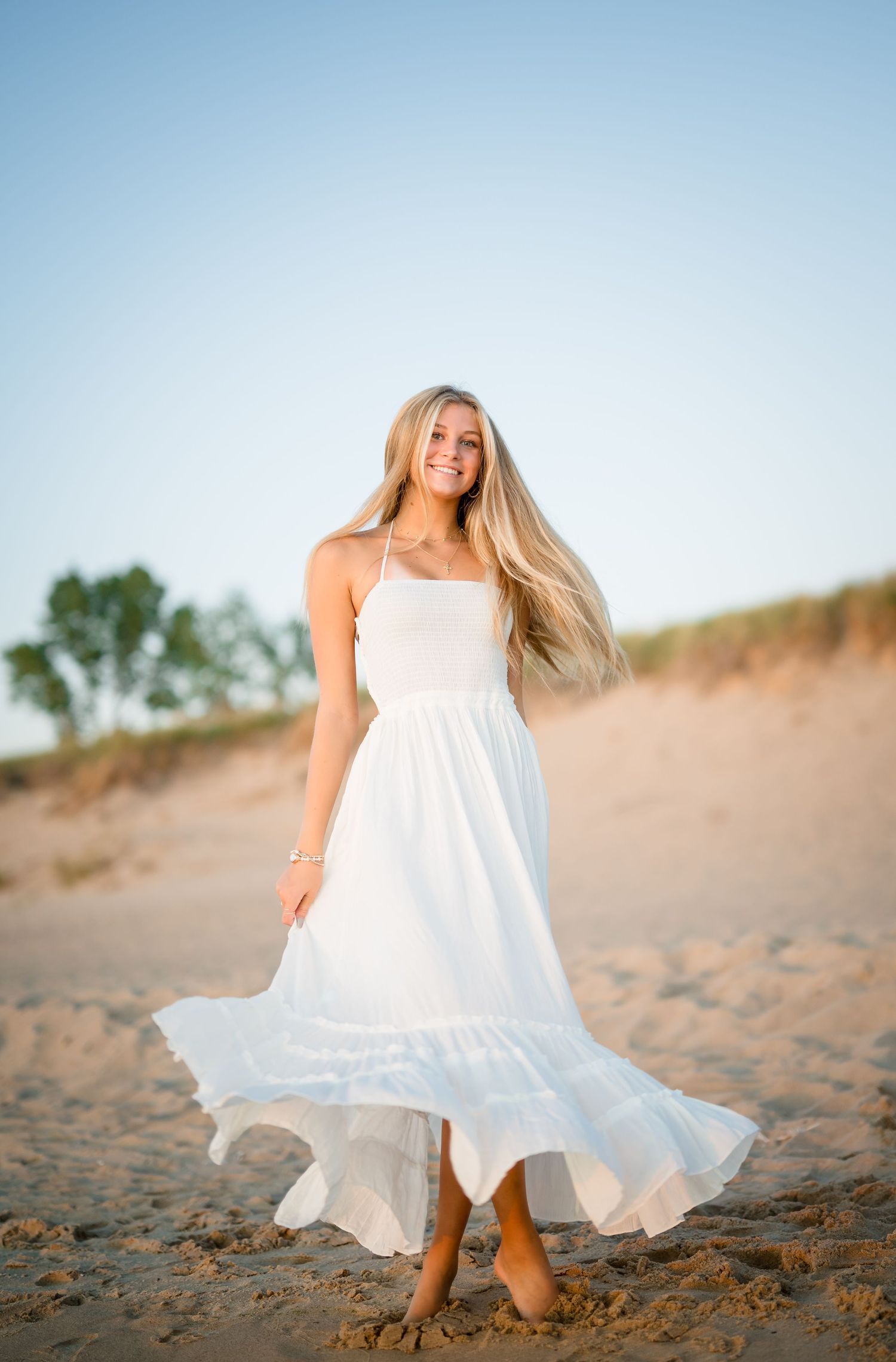 Senior girl in flowing white dress on sandy beach during golden hour photo session at Lake Michigan.