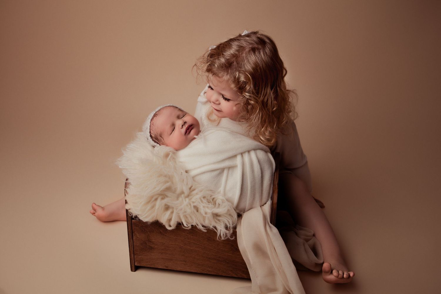 Newborn baby sleeping wrapped in a neutral blanket in a baby bed during a newborn photography session with big sister