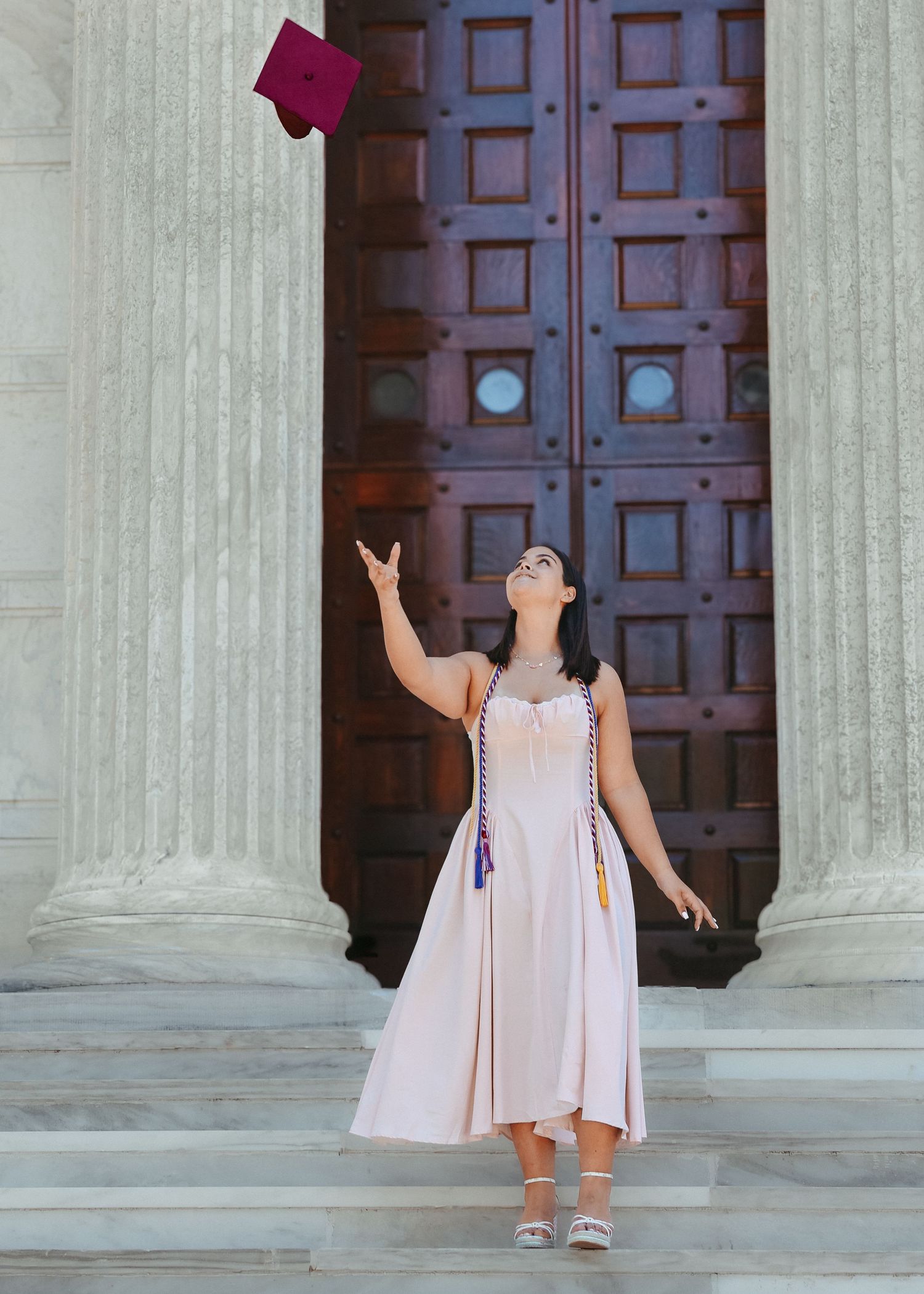 A high school graduate in a light pink dress smiling and holding a graduation pennant while posing next to a classic stone column at Princet