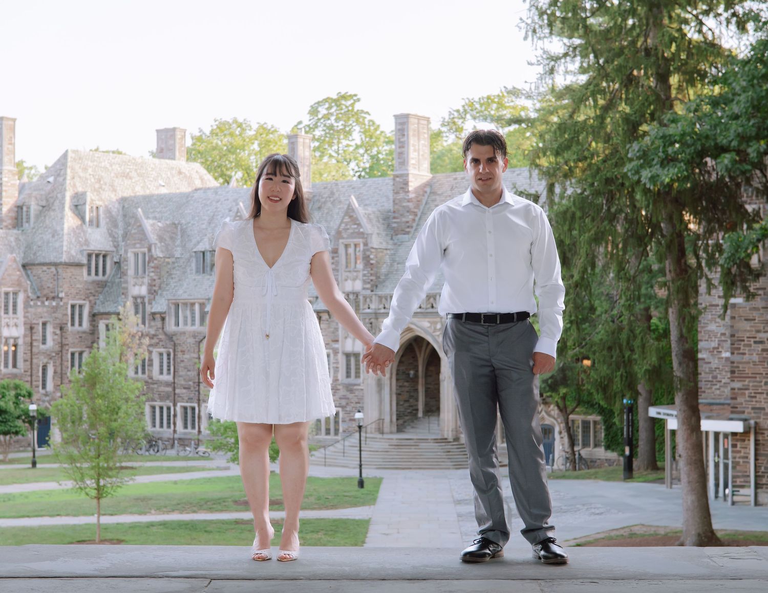 An engaged couple holding hands and walking along a stone path in front of a historic gothic-style building at Princeton University.
