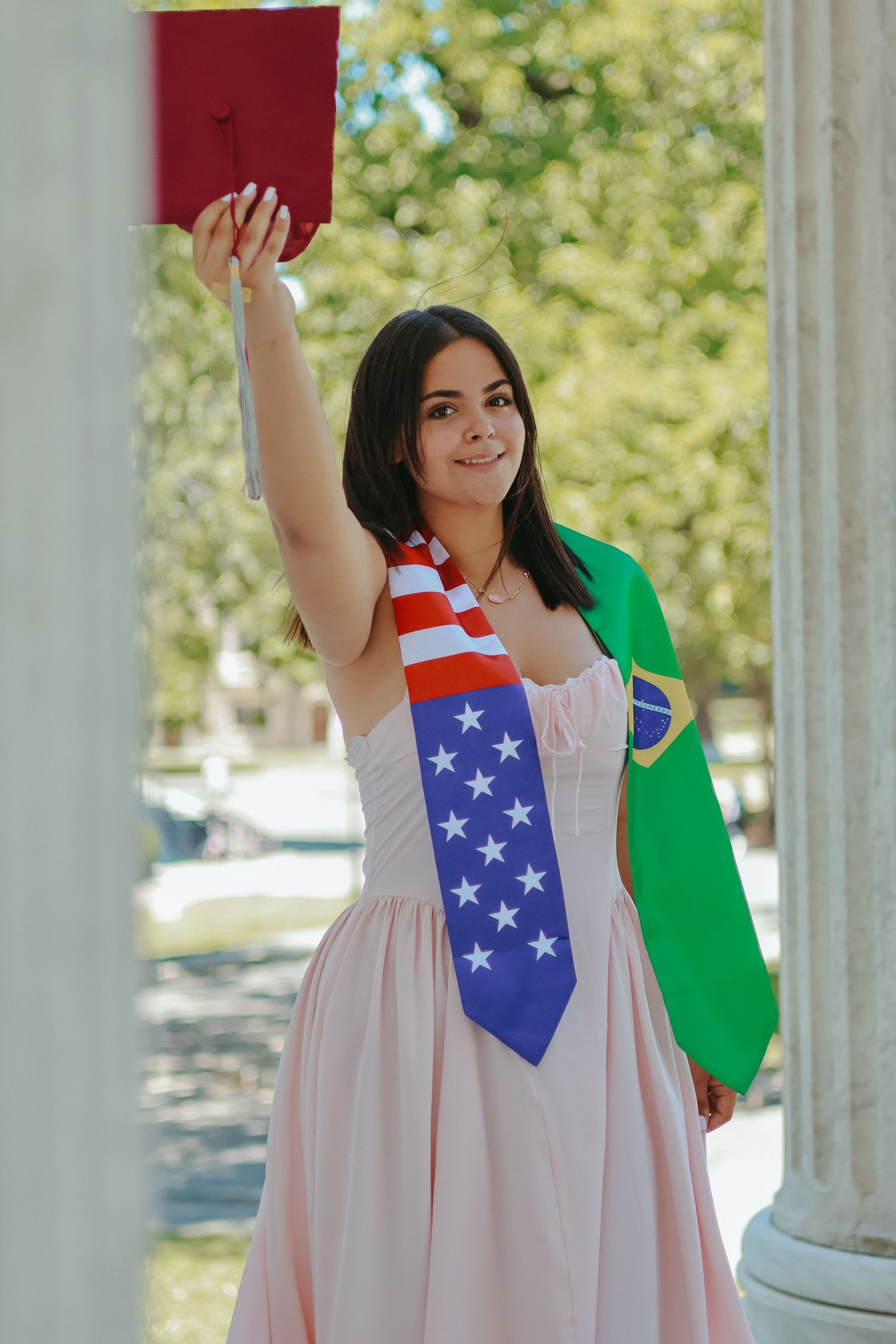 A high school graduate in a pink dress smiling and holding a graduation pennant while posing next to a classic stone column in Princeton