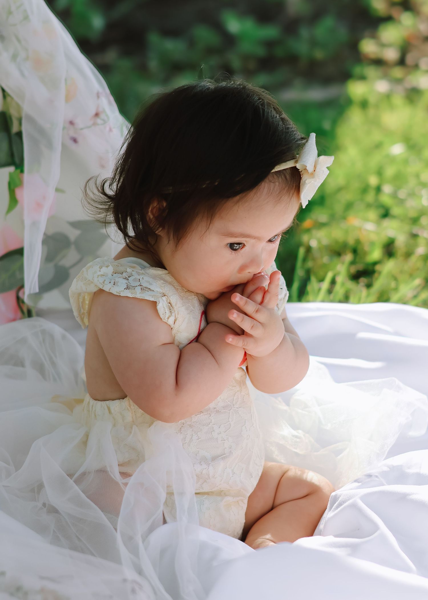A baby girl celebrating her first birthday, wearing a white tulle dress and sitting on a blanket outdoors in a sunlit grassy field.