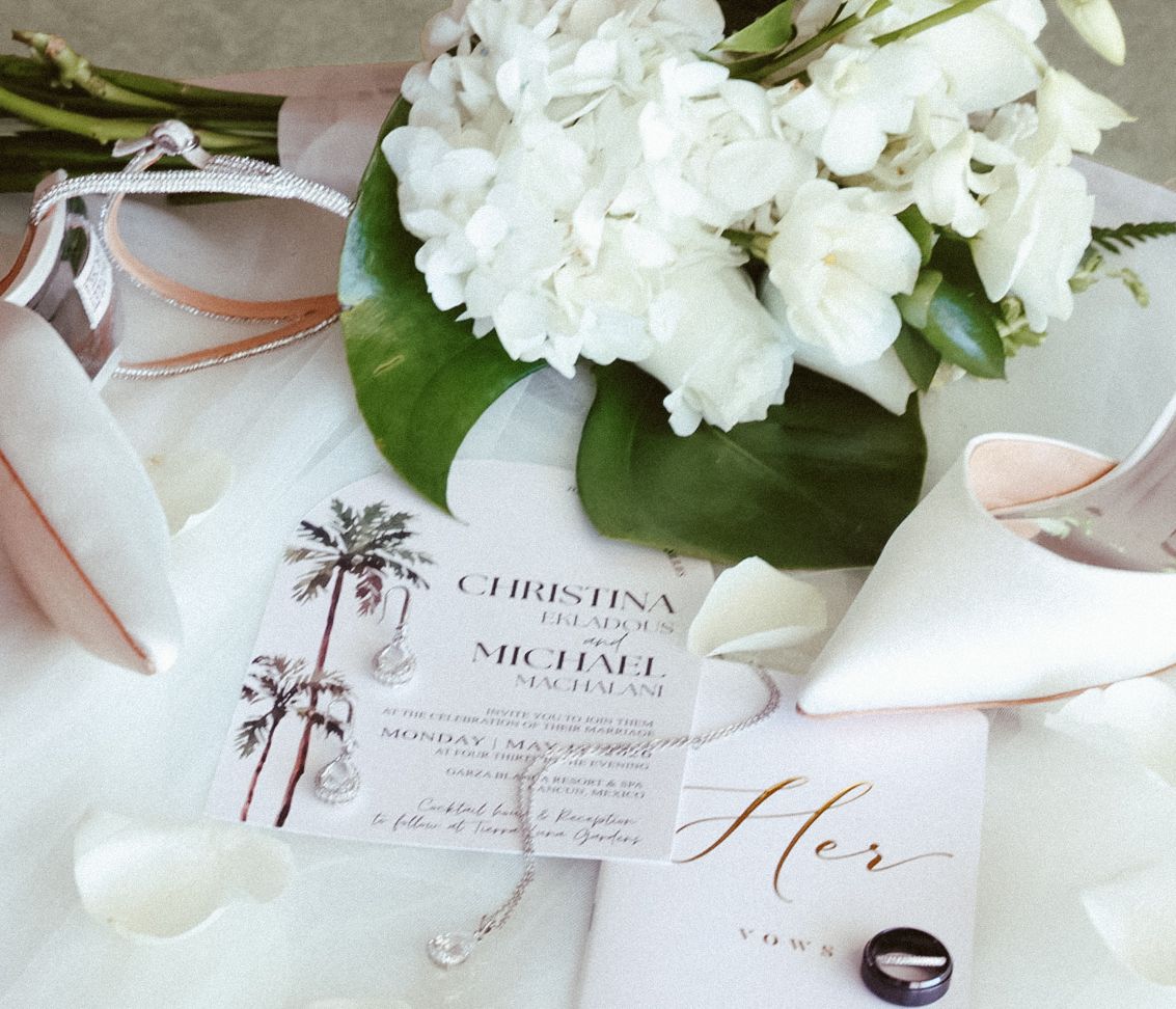 An elegant flat lay of destination wedding invitations, bridal perfume, and white floral details featuring a custom crest for a Mexico weddi
