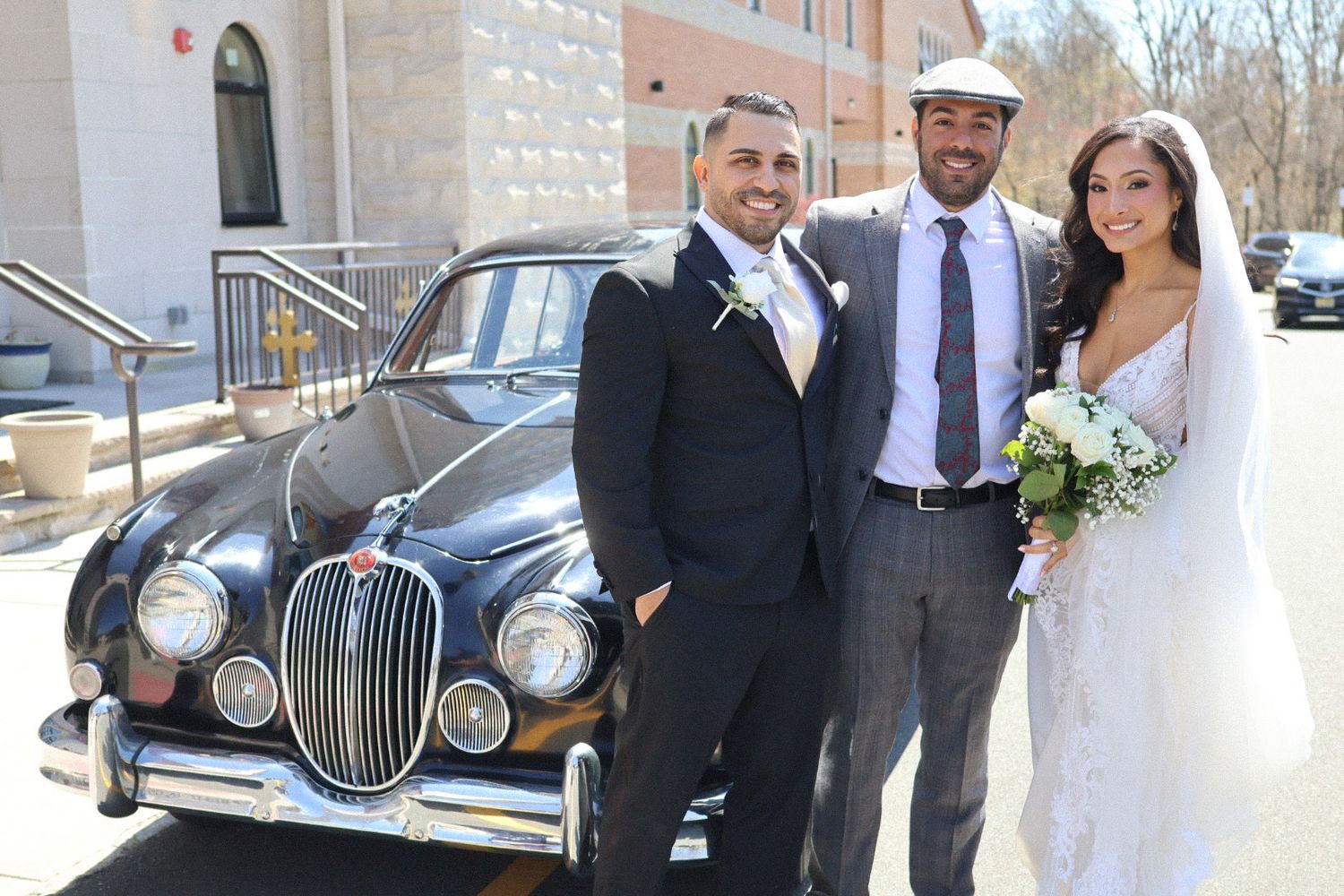 A bride and groom in elegant wedding attire posing with family, friends, and their chauffeur next to a classic, vintage black car outdoors a