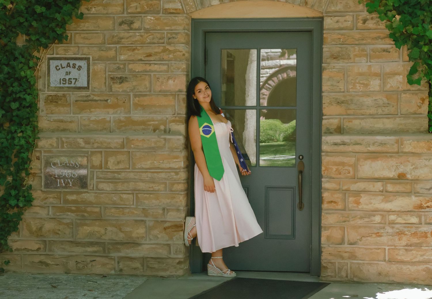 A high school graduate in a pink dress smiling and holding a graduation pennant while posing next to a classic stone column in Princeton