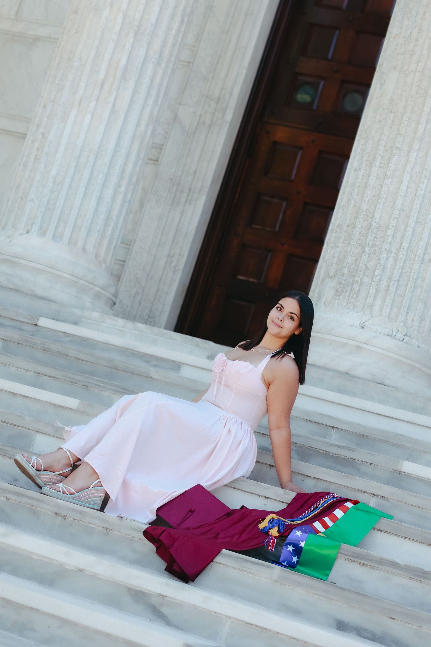 A high school graduate in a pink dress smiling and holding a graduation pennant while posing next to a classic stone column in Princeton