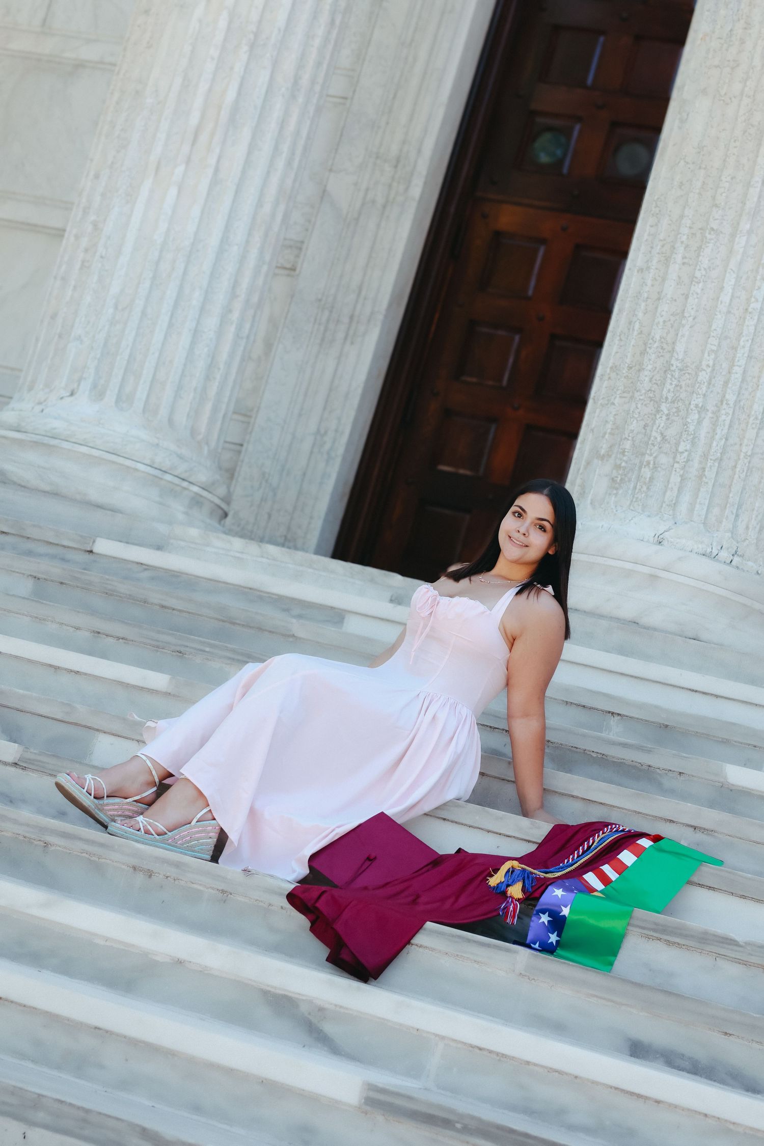 A high school graduate in a light pink dress smiling and holding a graduation pennant while posing next to a classic stone column at Princet