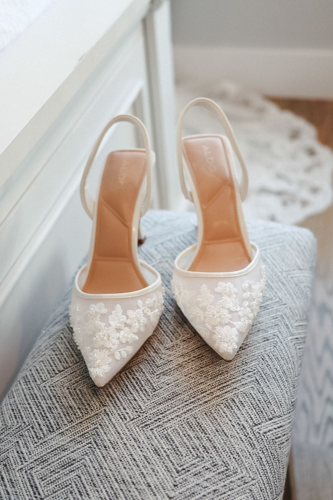 A close-up shot of elegant white lace bridal heels resting on a stylized mat by a sunlit window at a destination wedding venue.