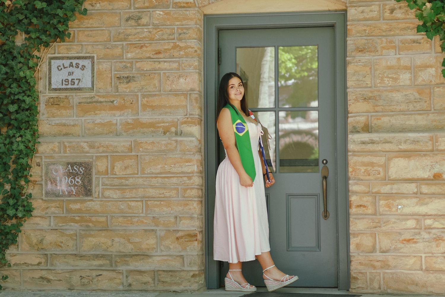 A high school graduate in a pink dress smiling and holding a graduation pennant while posing next to a classic stone column in Princeton