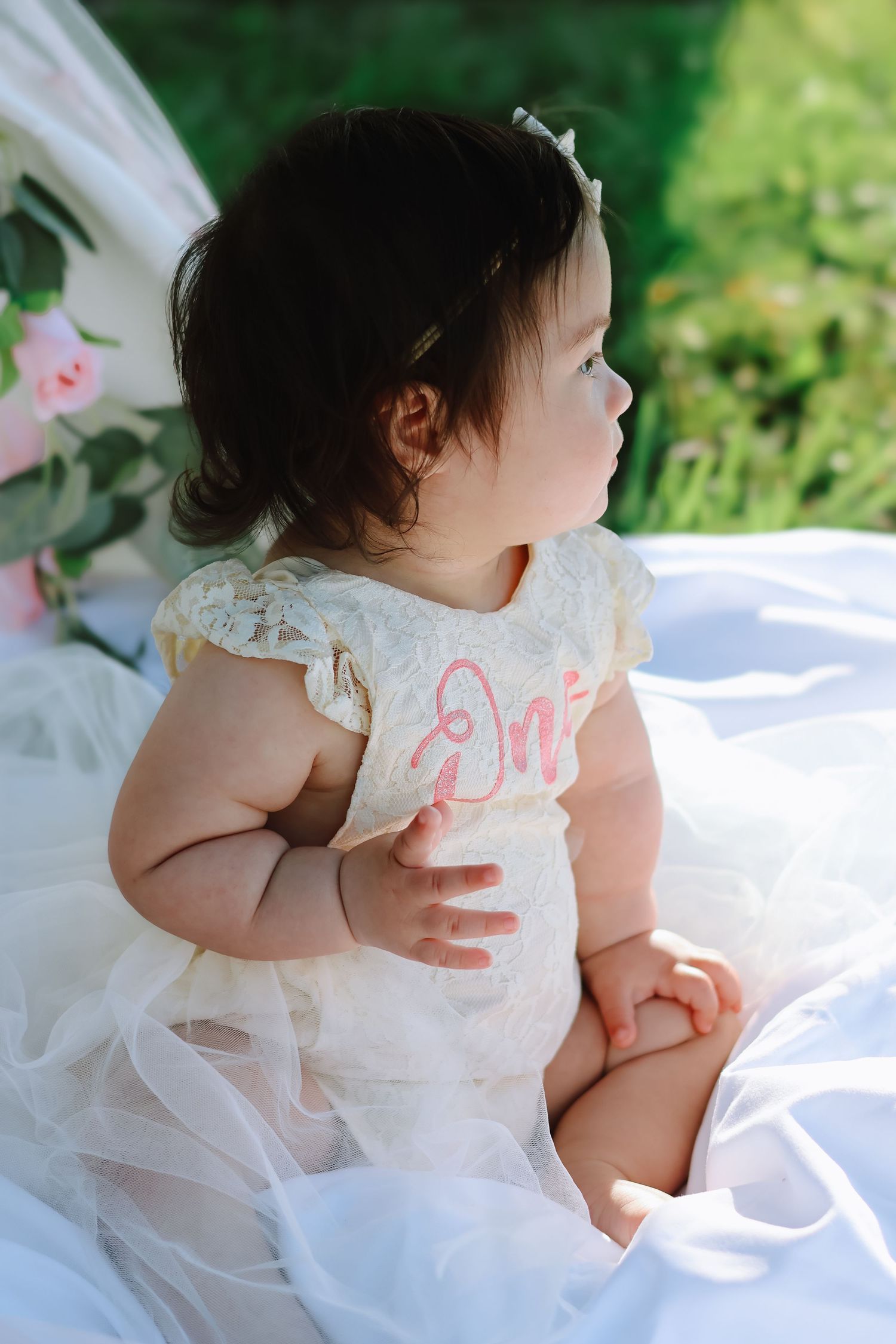 A baby girl celebrating her first birthday, wearing a white tulle dress and sitting on a blanket outdoors in a sunlit grassy field.