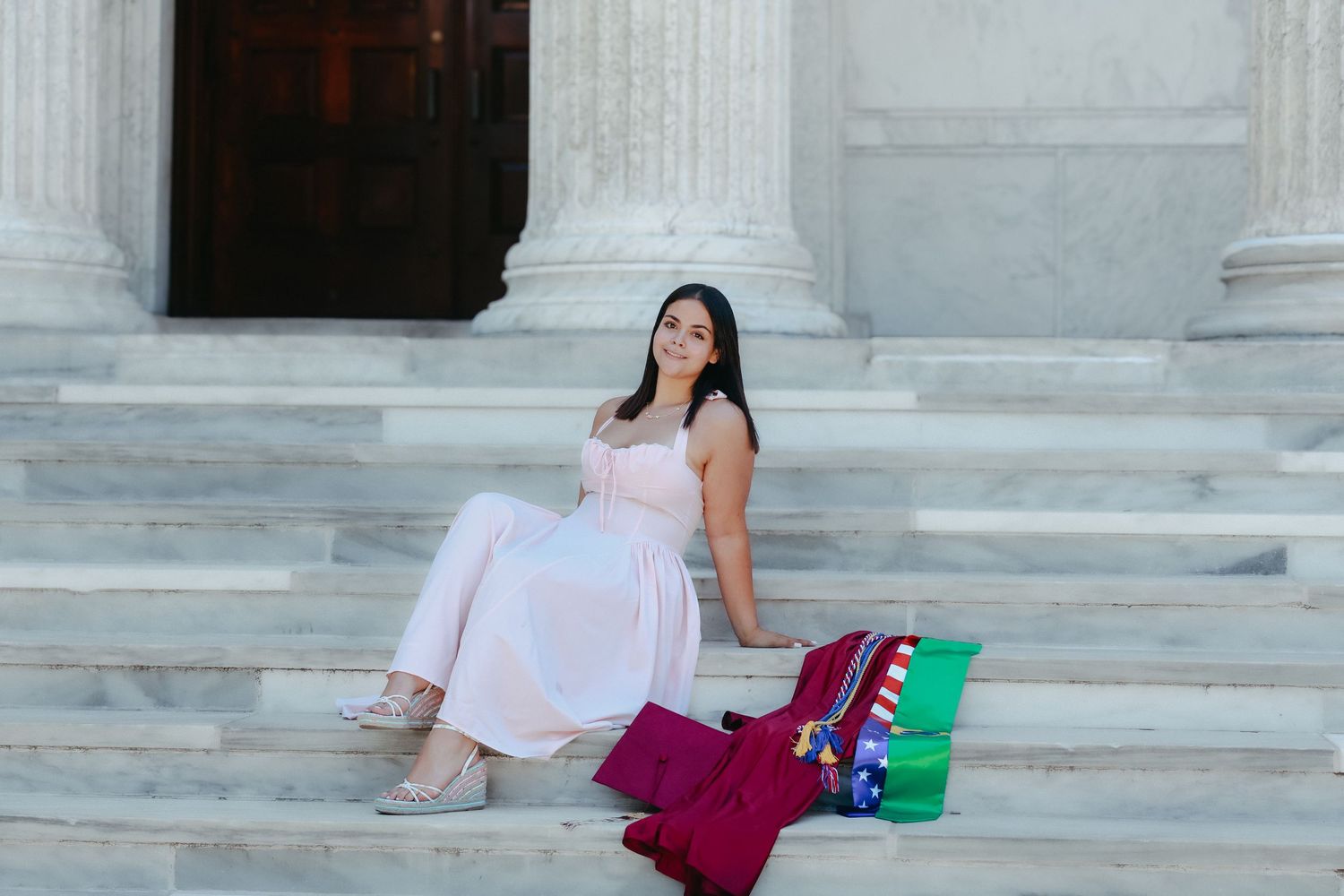 A high school graduate in a pink dress smiling and holding a graduation pennant while posing next to a classic stone column in Princeton