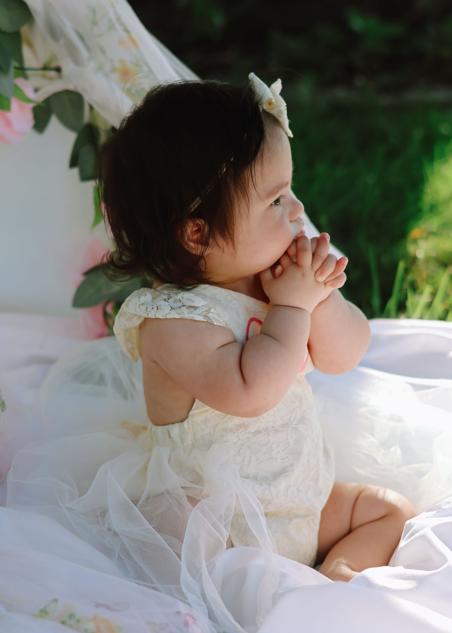 A baby girl celebrating her first birthday, wearing a white tulle dress and sitting on a blanket outdoors in a sunlit grassy field.