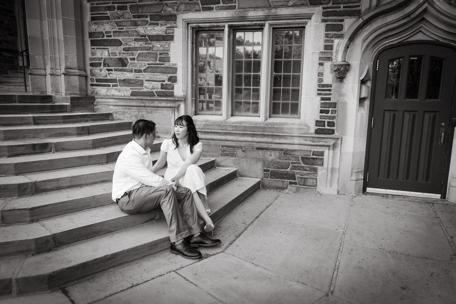 An engaged couple holding hands and walking along a stone path in front of a historic gothic-style building at Princeton University.