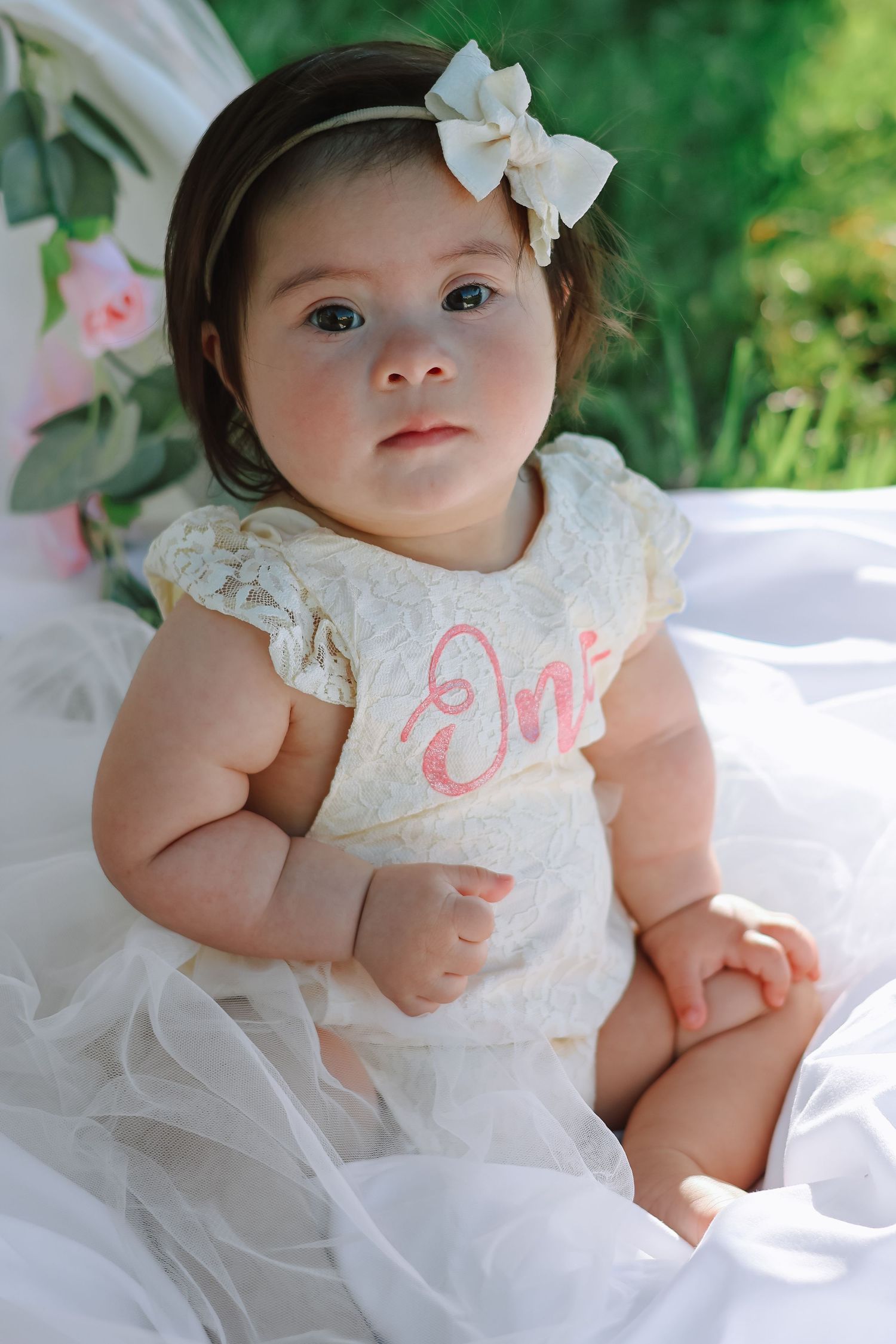 A baby girl celebrating her first birthday, wearing a white tulle dress and sitting on a blanket outdoors in a sunlit grassy field.