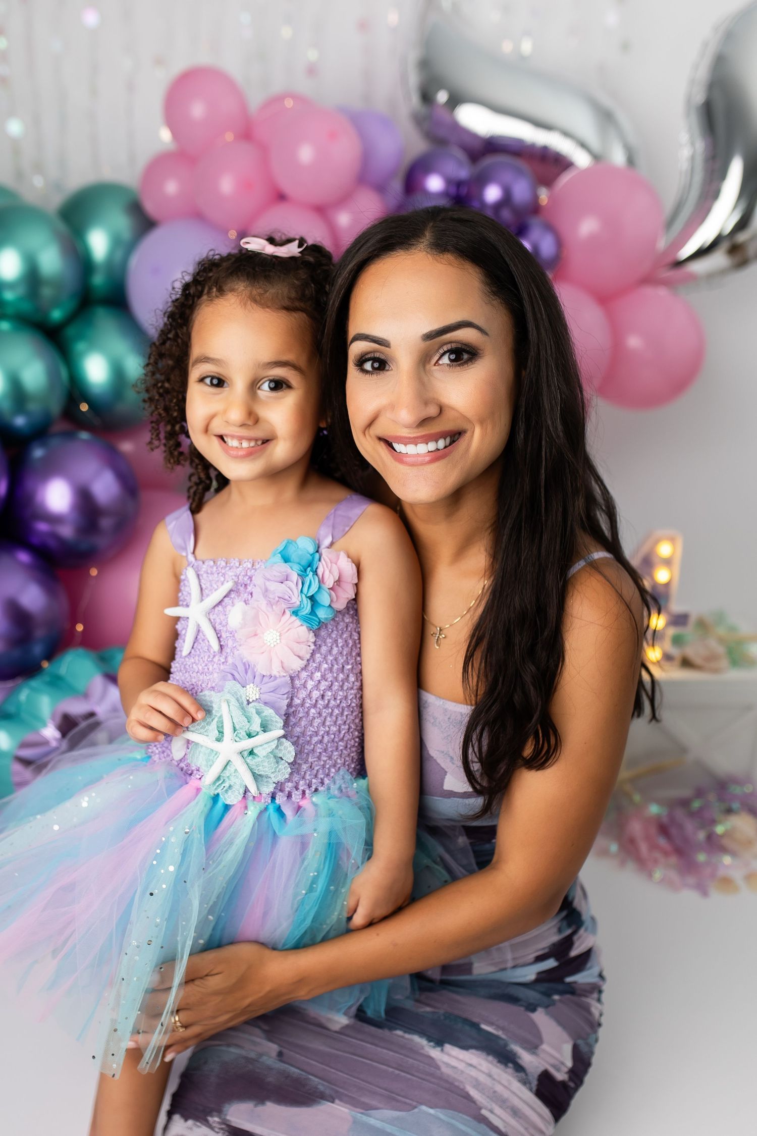 A smiling mother and her young daughter posing together in front of a vibrant pink, purple, and green balloon arch during a family portrait