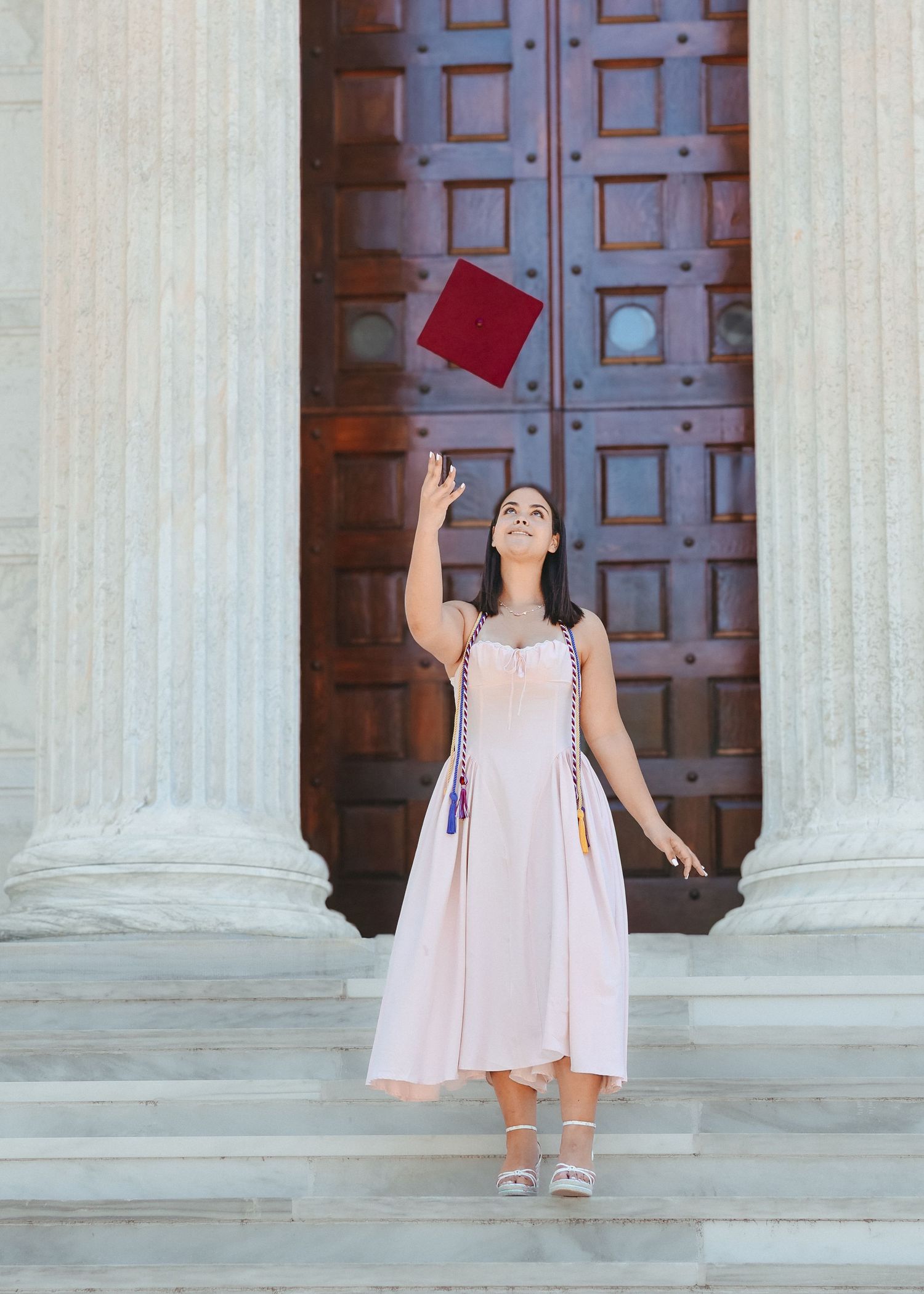 A high school graduate in a pink dress smiling and holding a graduation pennant while posing next to a classic stone column in Princeton